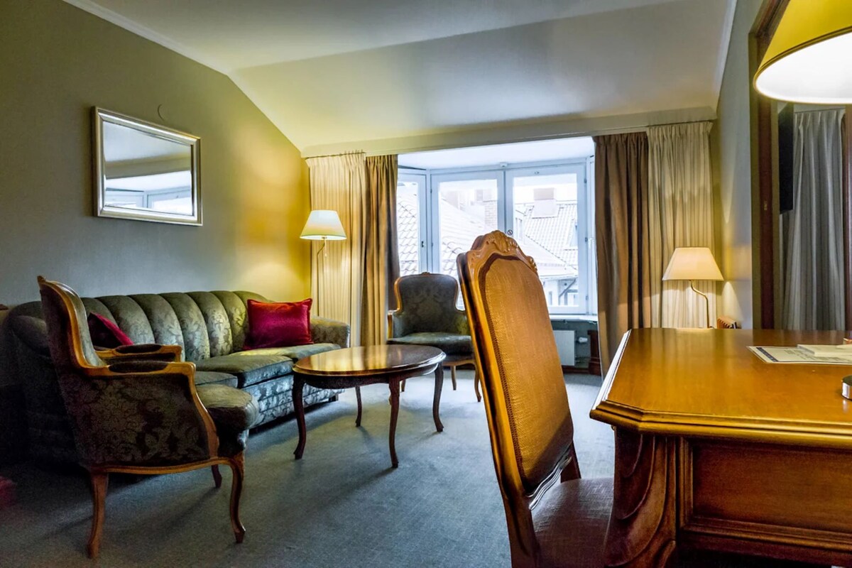 Habitación de hotel con sofá, sillones, una mesa de comedor y un amplio ventanal con cortinas.