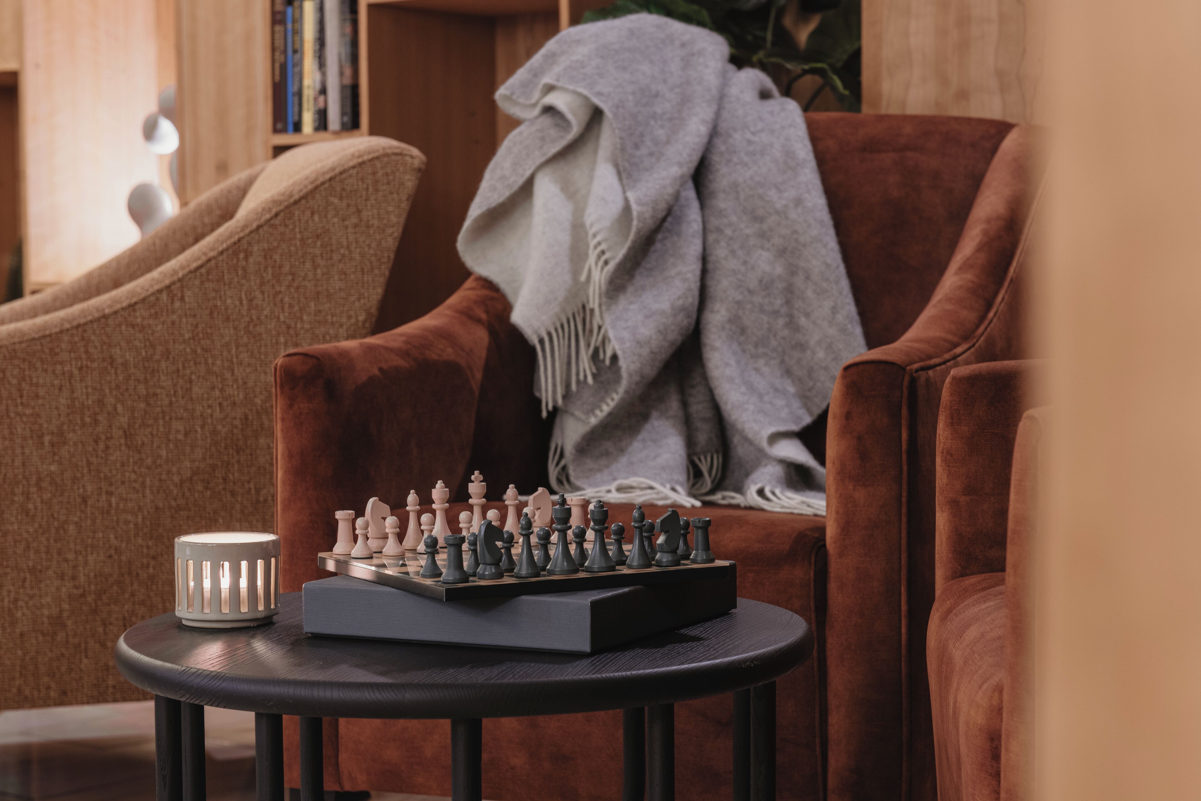 A chessboard on a small table in a cozy room with a brown armchair and a bookshelf in the background.