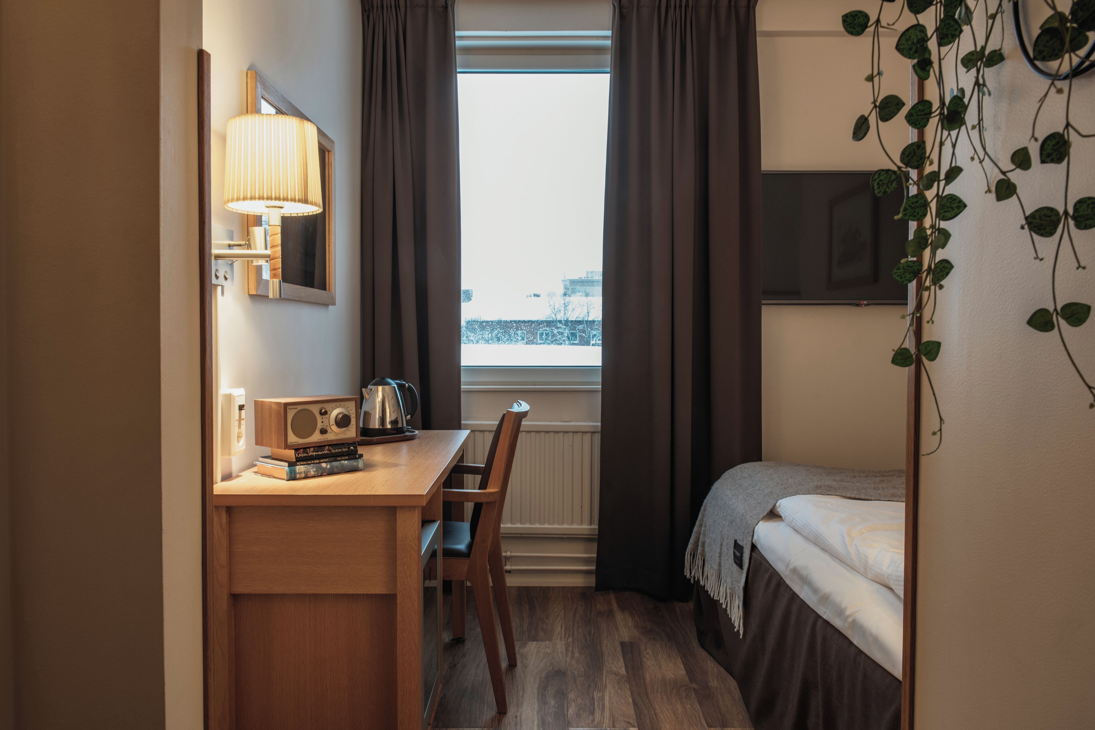 Habitación de hotel pequeña con escritorio de madera, una silla, una cama, ventana con cortinas, lámpara de pared y radiador.