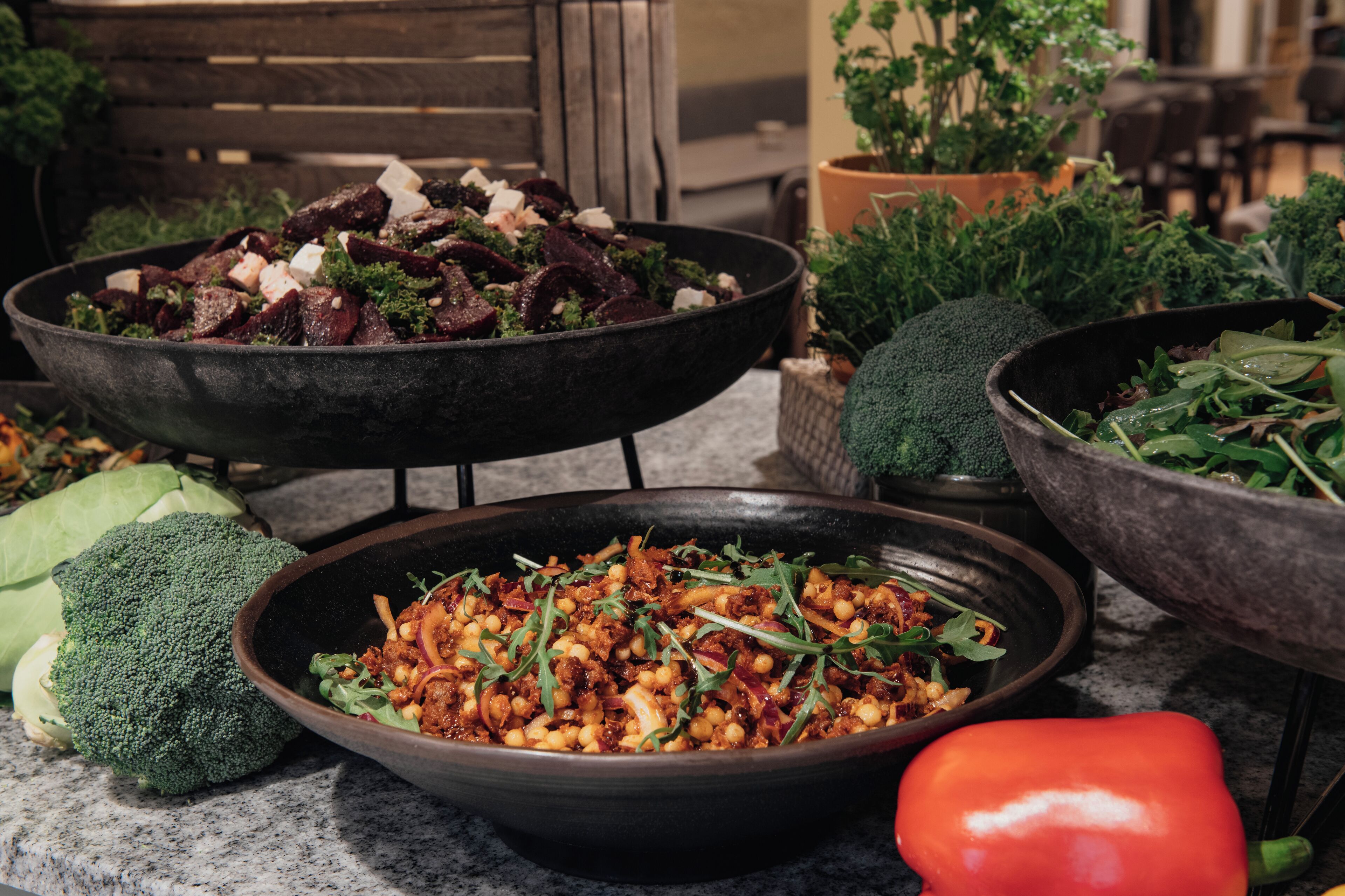 On trouve une variété de plats servis dans de grands bols, notamment une salade composée de légumes verts et de viande, un plat à base de pois chiches et de légumes, et un bol contenant un poivron rouge.