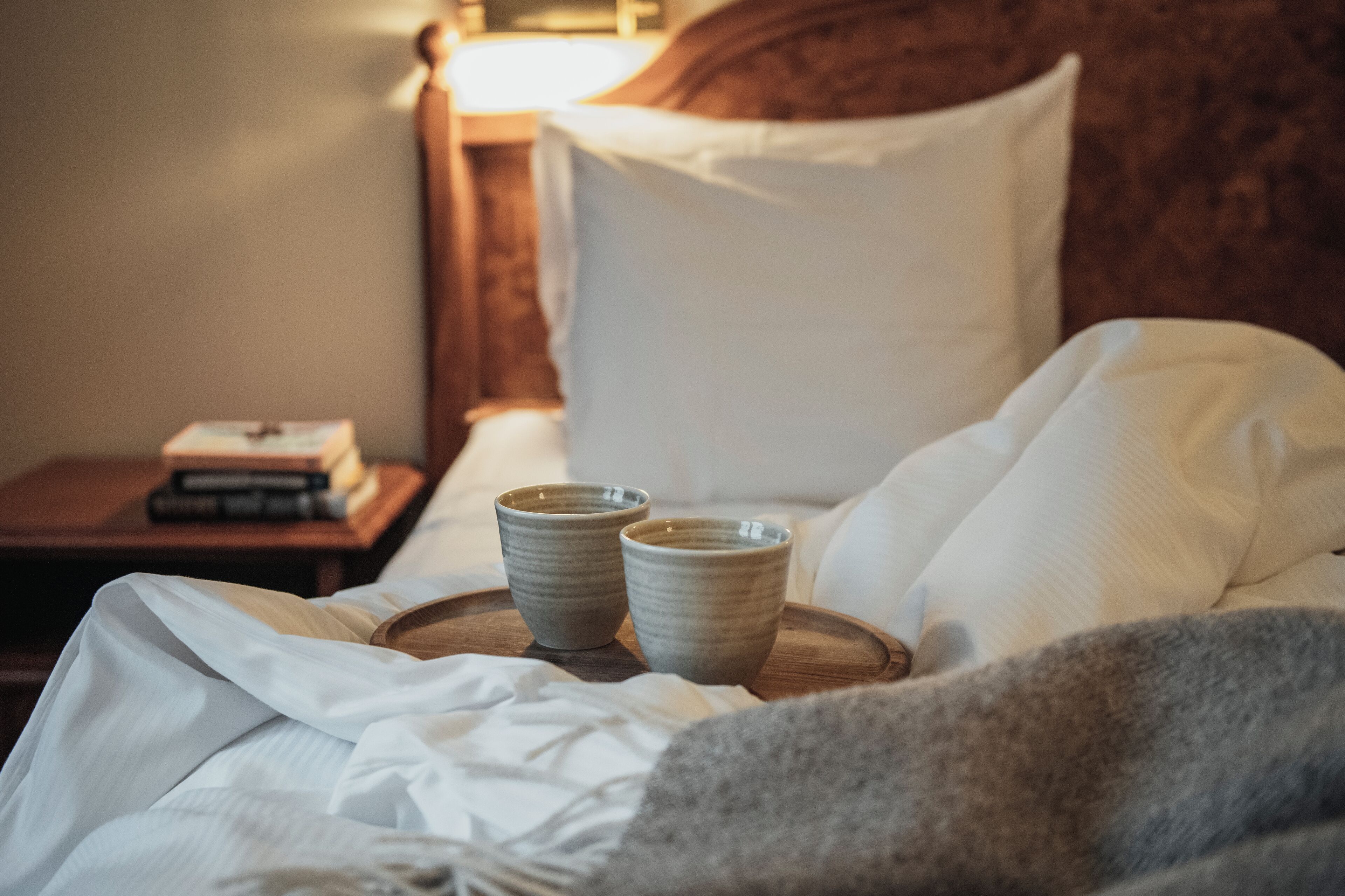 A bed with white linens, a wooden tray with two ceramic cups, and a nightstand with books and a lamp.