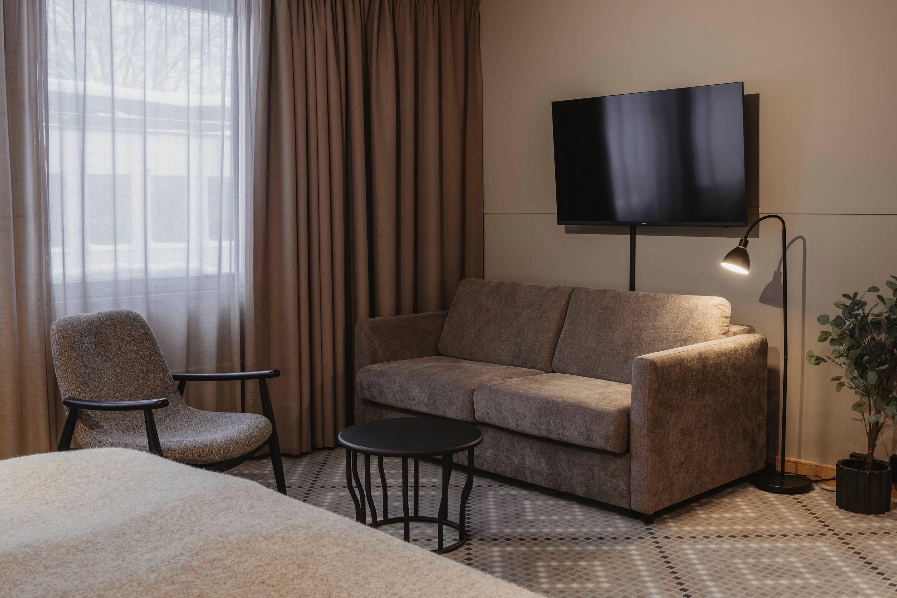 A hotel room with a sofa, a chair, a small table, a TV mounted on the wall, and a potted plant.