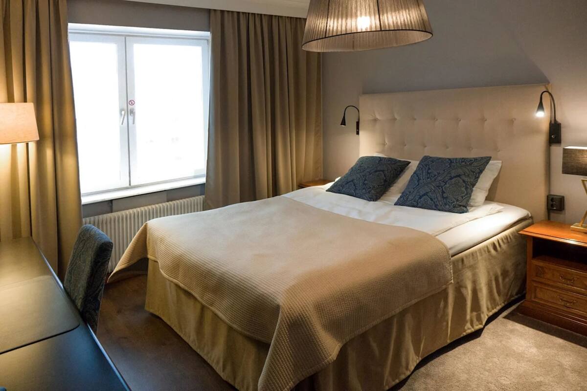 A hotel room with a bed, a desk, a chair, a window with curtains, and a lamp.
