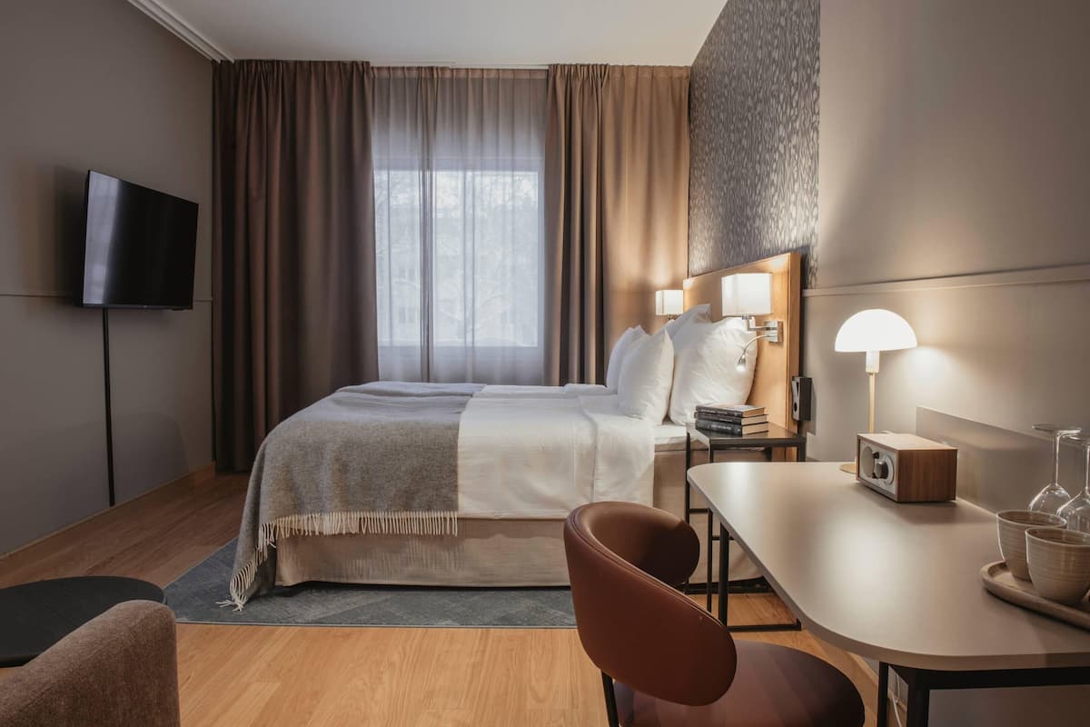 A hotel room with a bed, bedside lamps, a flat-screen TV, a desk with a lamp, and a chair.