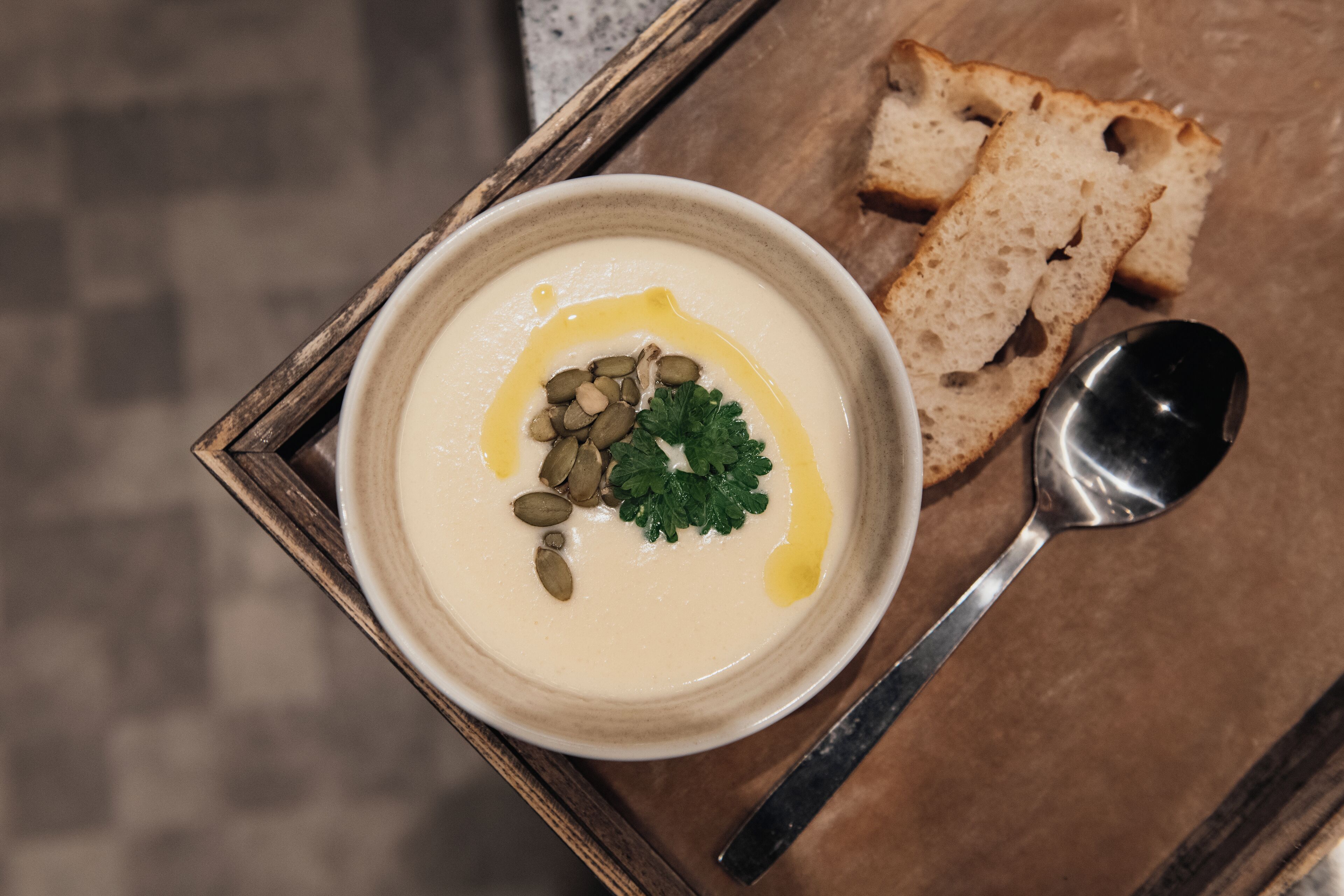 A bowl of creamy soup garnished with seeds and parsley, accompanied by slices of bread and a spoon on a wooden tray.