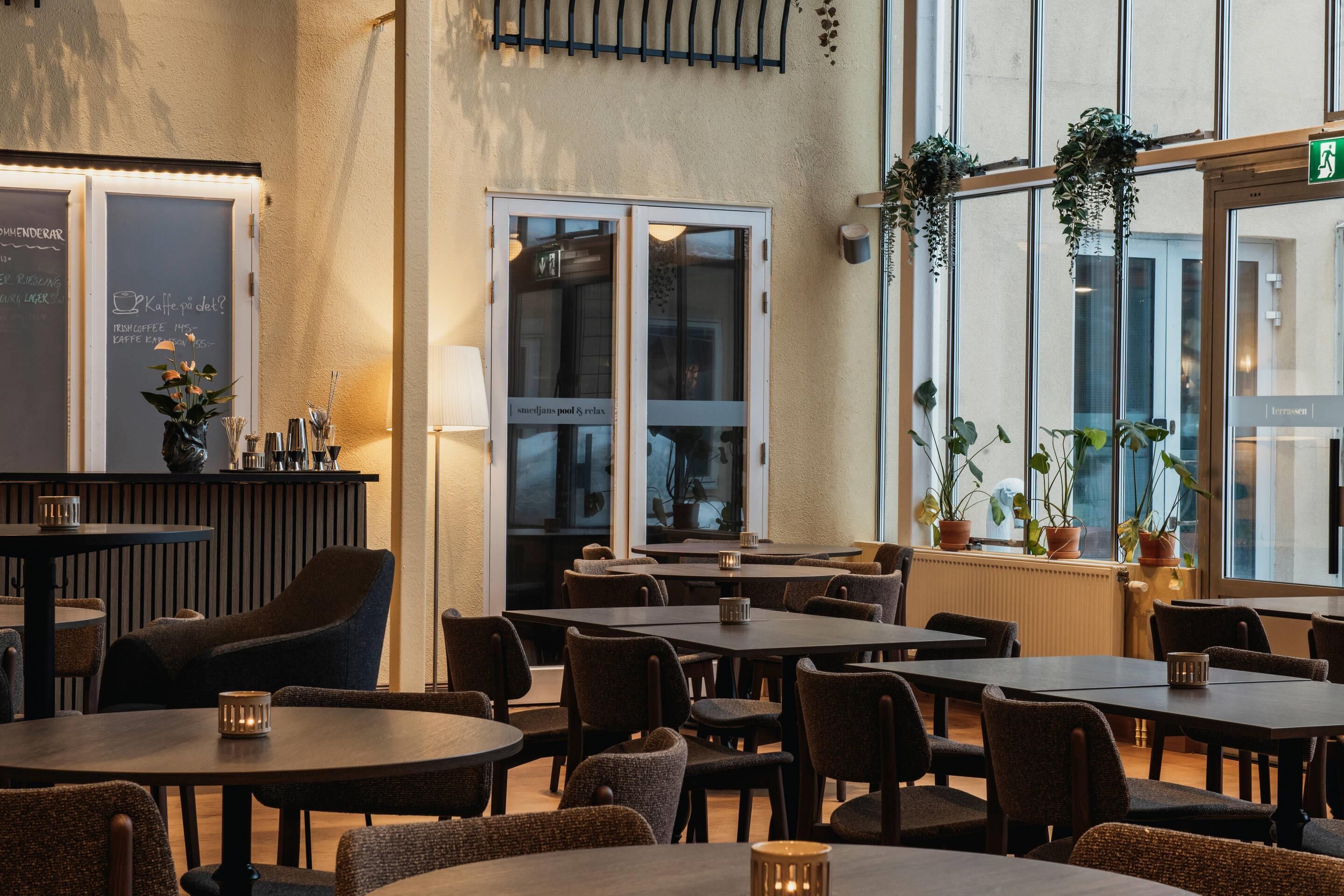 Un restaurante con mesas y sillas, una zona de bar y amplios ventanales con plantas.