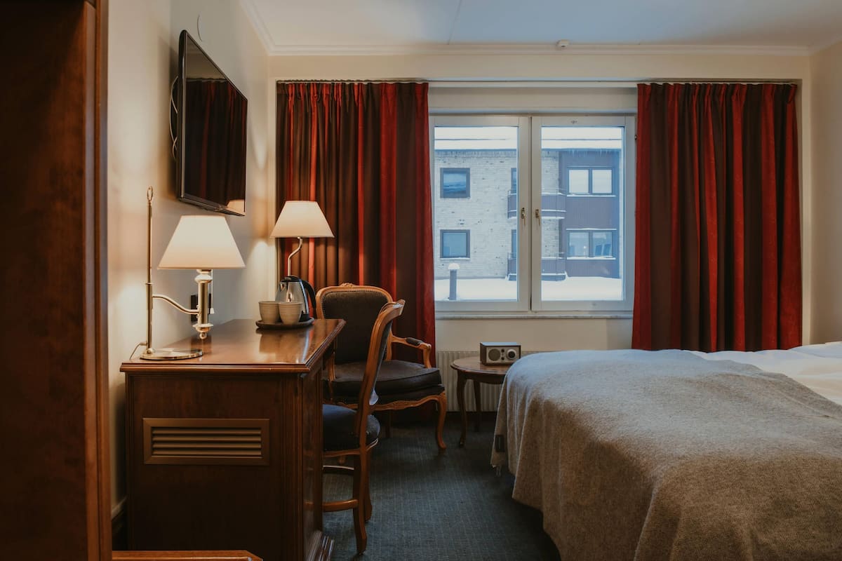 A hotel room with a bed, a desk with a lamp, a chair, a radio, and a window with curtains.