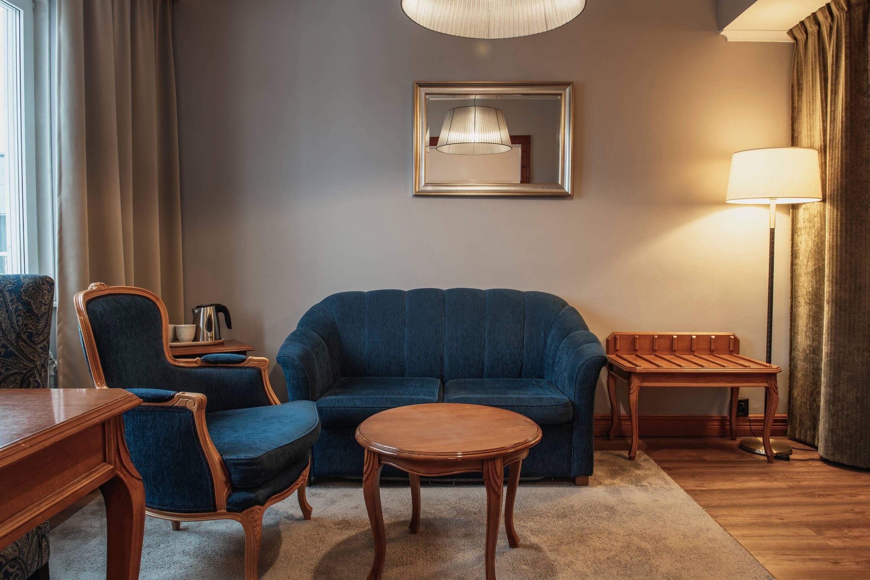 Ein Zimmer mit einem blauen Sofa, einem Holztisch, einem Nachttisch, einem Sessel, einer Lampe und einem gerahmten Spiegel an der Wand.