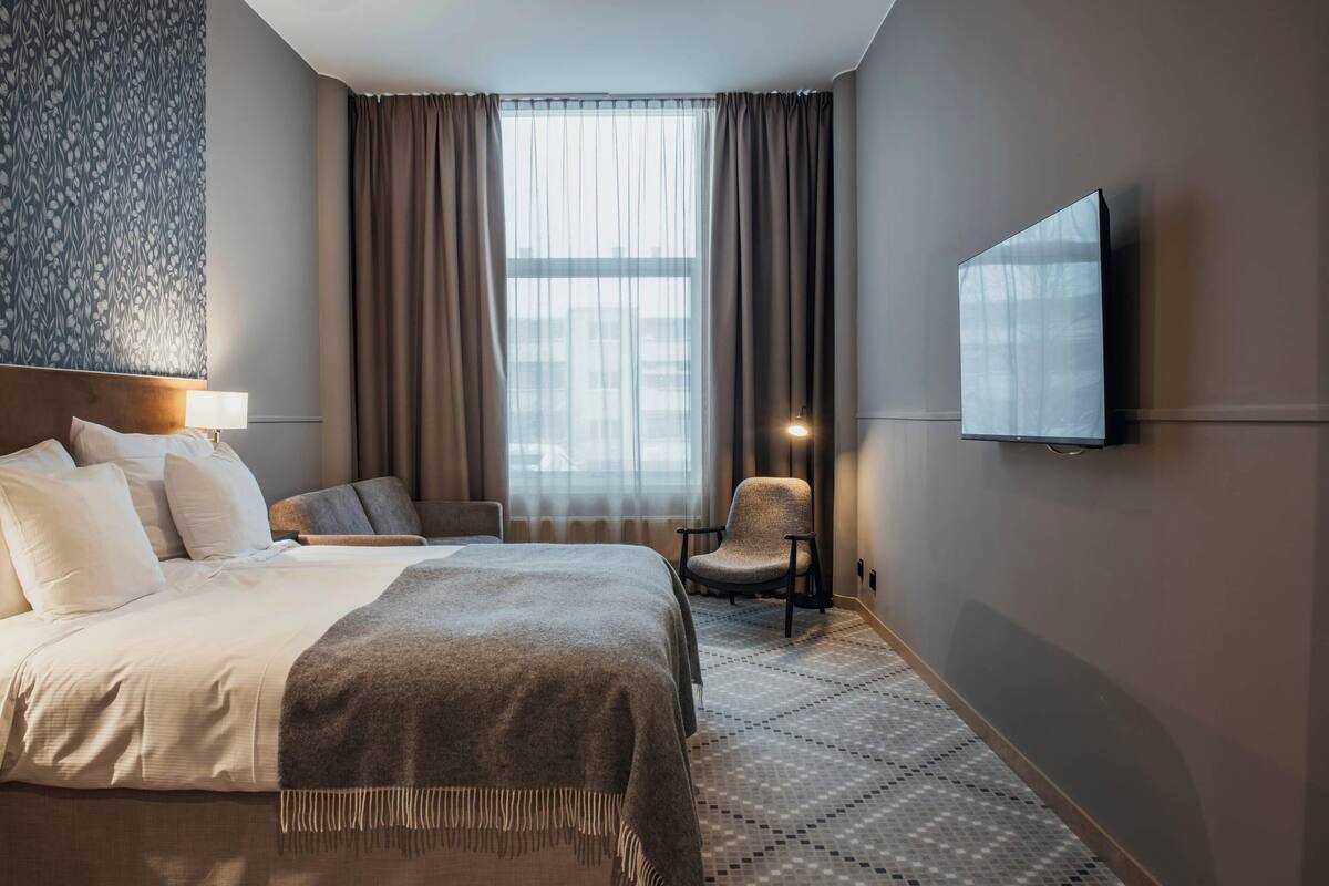 A hotel room with a bed, a chair, a television, and a large window with curtains.