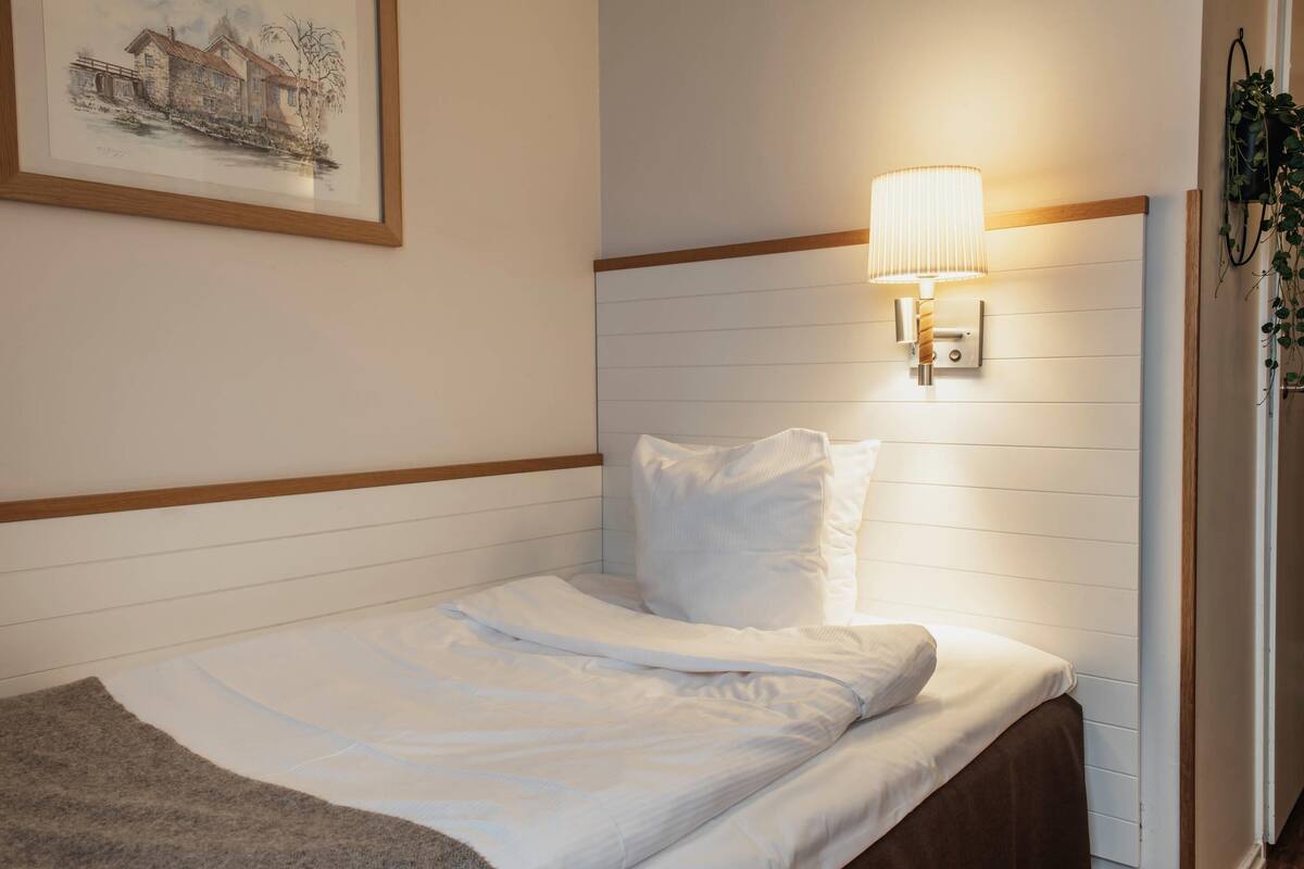 A neatly made bed with white linens, a wall-mounted lamp, and a framed picture of a building.