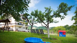 A grassy area with two blue play structures, a red and blue playhouse, and a building with balconies and a view of hills. - Pensiunea Toscana (Brasov)