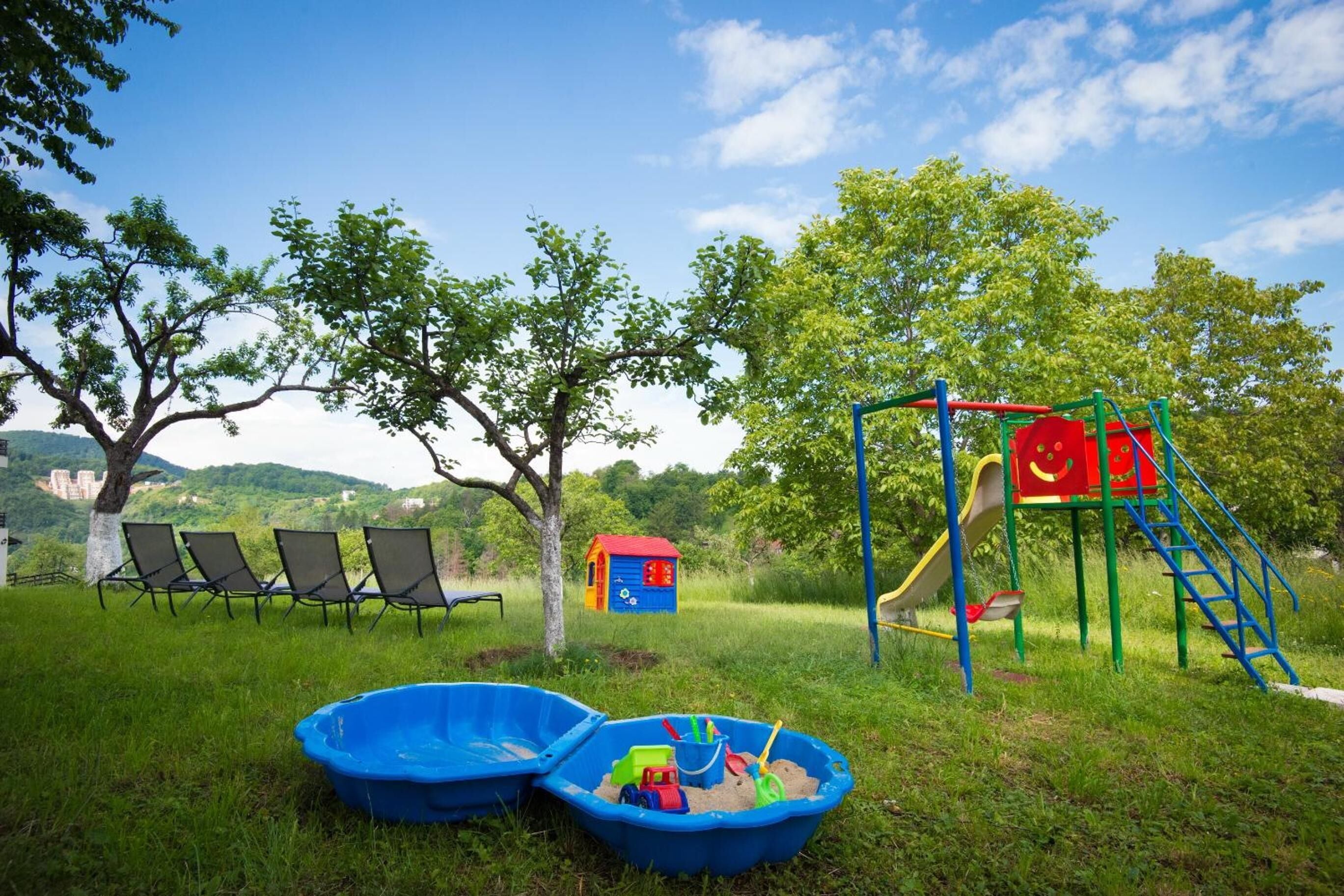 Un parque infantil con tobogán, columpios y una casita. También hay dos piscinas de plástico azules con juguetes y una fila de sillas.