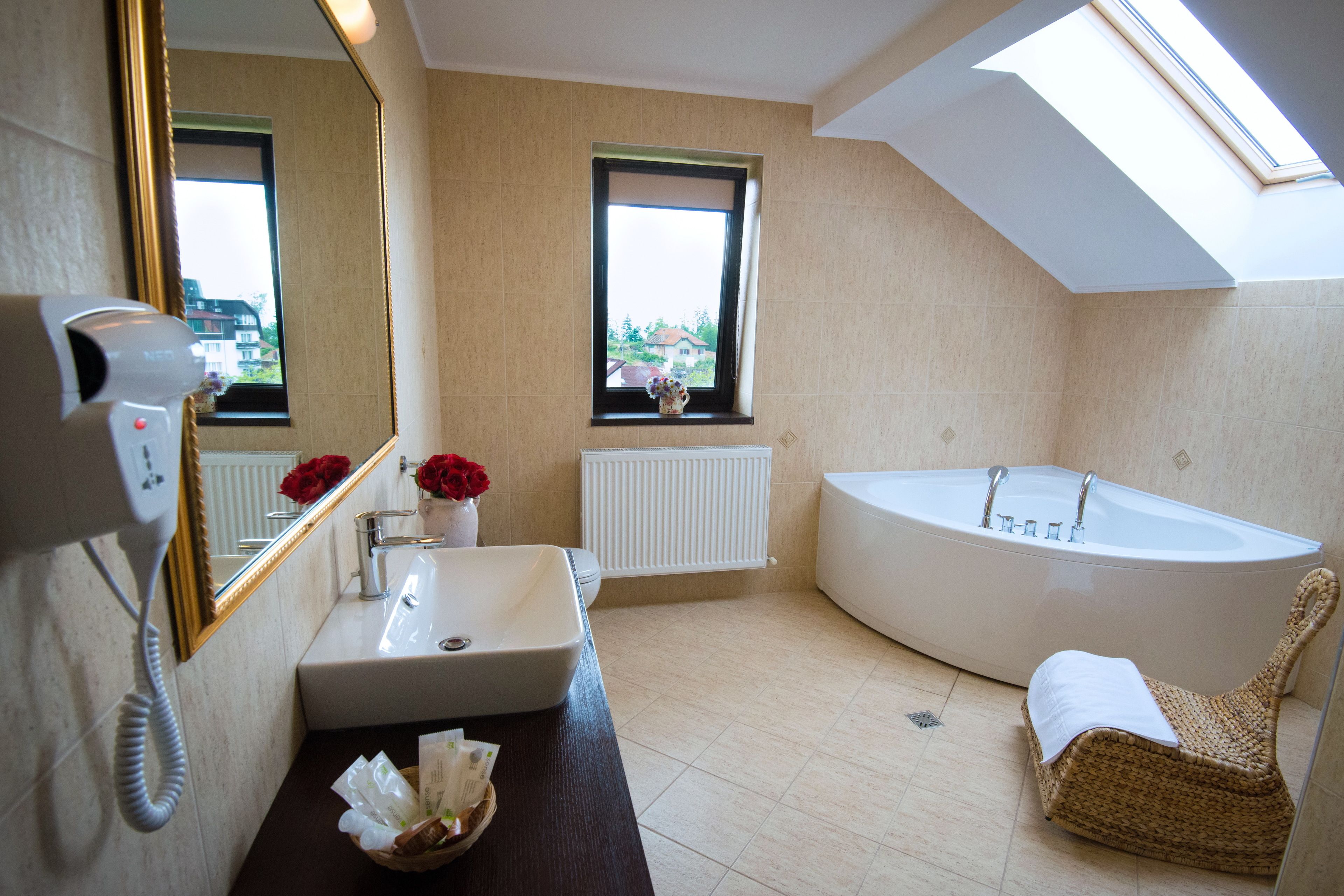 A modern bathroom with a large bathtub, a sink with a vase of red flowers, a wicker chair, and a skylight.