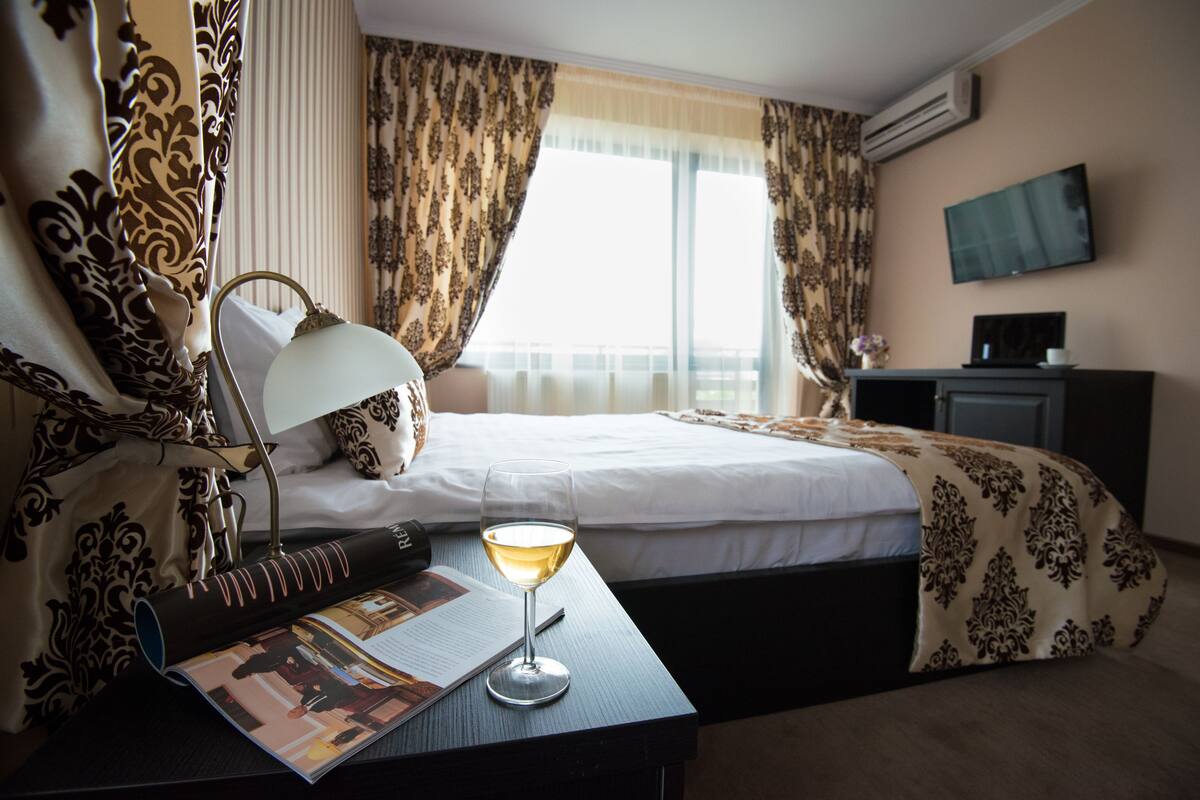 Een hotelkamer met een bed, een bureau met lamp, een glas wijn en een televisie aan de muur.