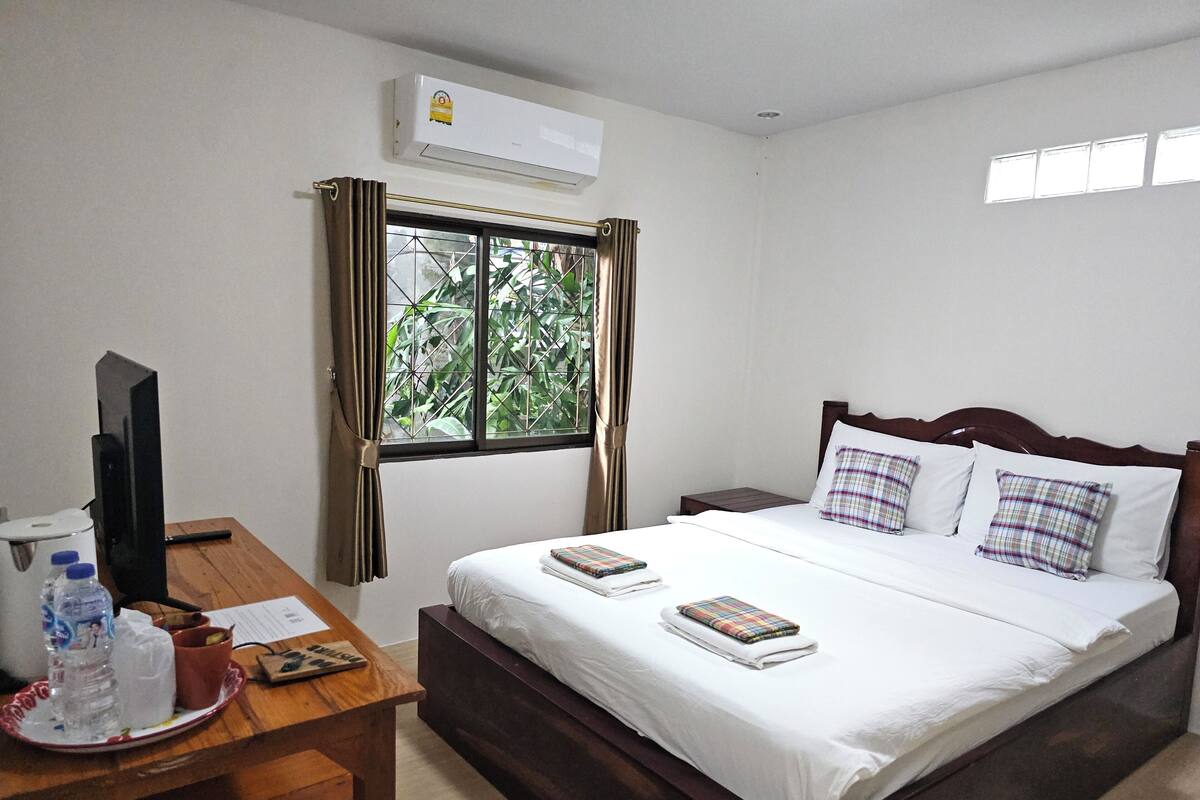 A hotel room with a bed, a desk with a computer, a window with curtains, and an air conditioning unit.