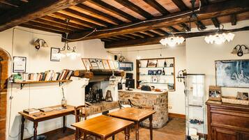 Un intérieur rustique avec des poutres apparentes, une cheminée en briques, des tables et des chaises en bois, ainsi que des étagères remplies de livres et d’objets décoratifs.