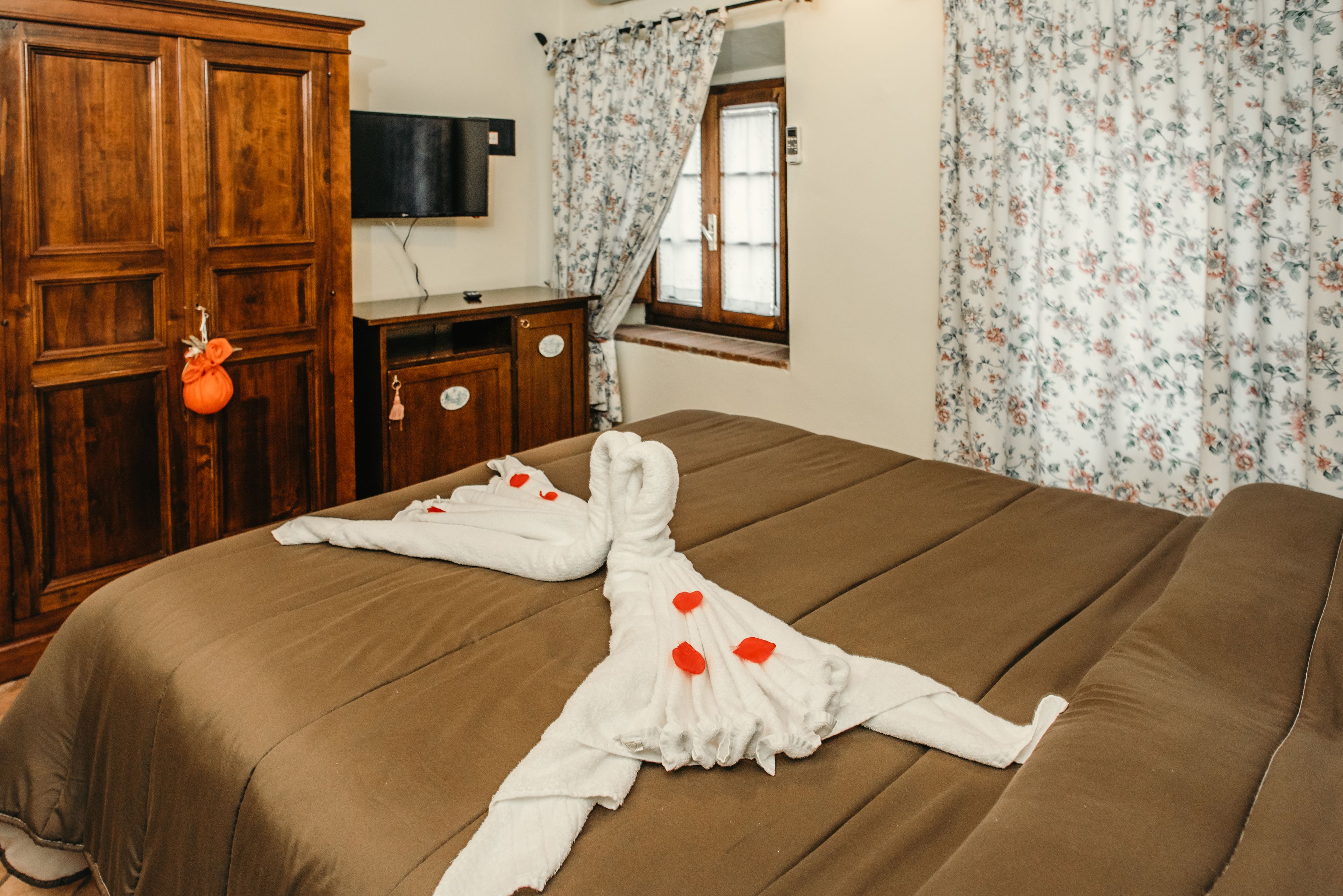 Een net opgemaakt bed met een handdoek en rozenblaadjes, een houten garderobe, een klein bureau en een raam met bloemen gordijnen.