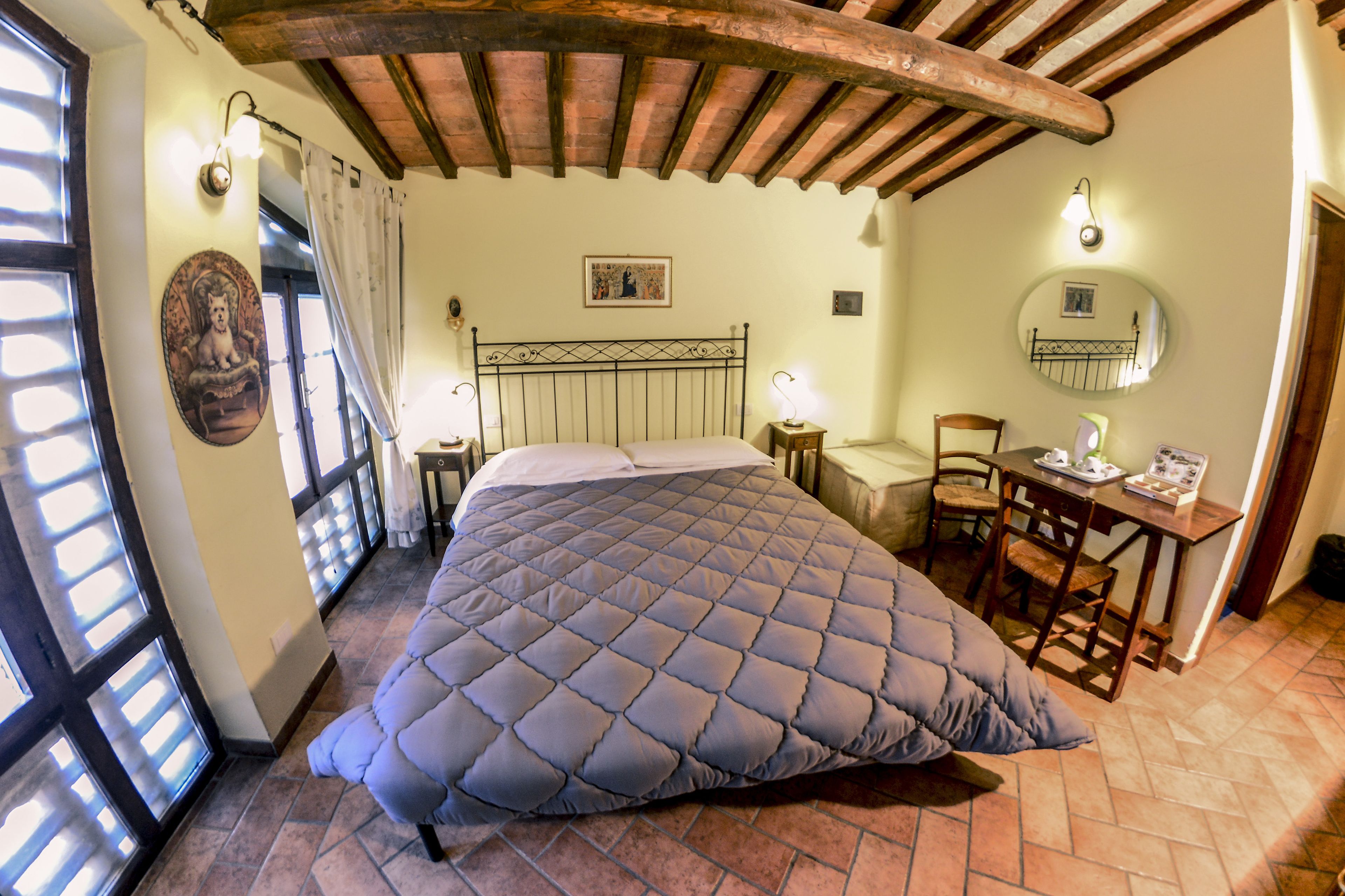 Un dormitorio con cama, escritorio y silla. Se ve una ventana con cortinas, un espejo fijado a la pared y un cuadro.