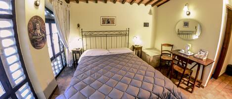 Un dormitorio con cama, escritorio y silla. Se ve una ventana con cortinas, un espejo fijado a la pared y un cuadro.