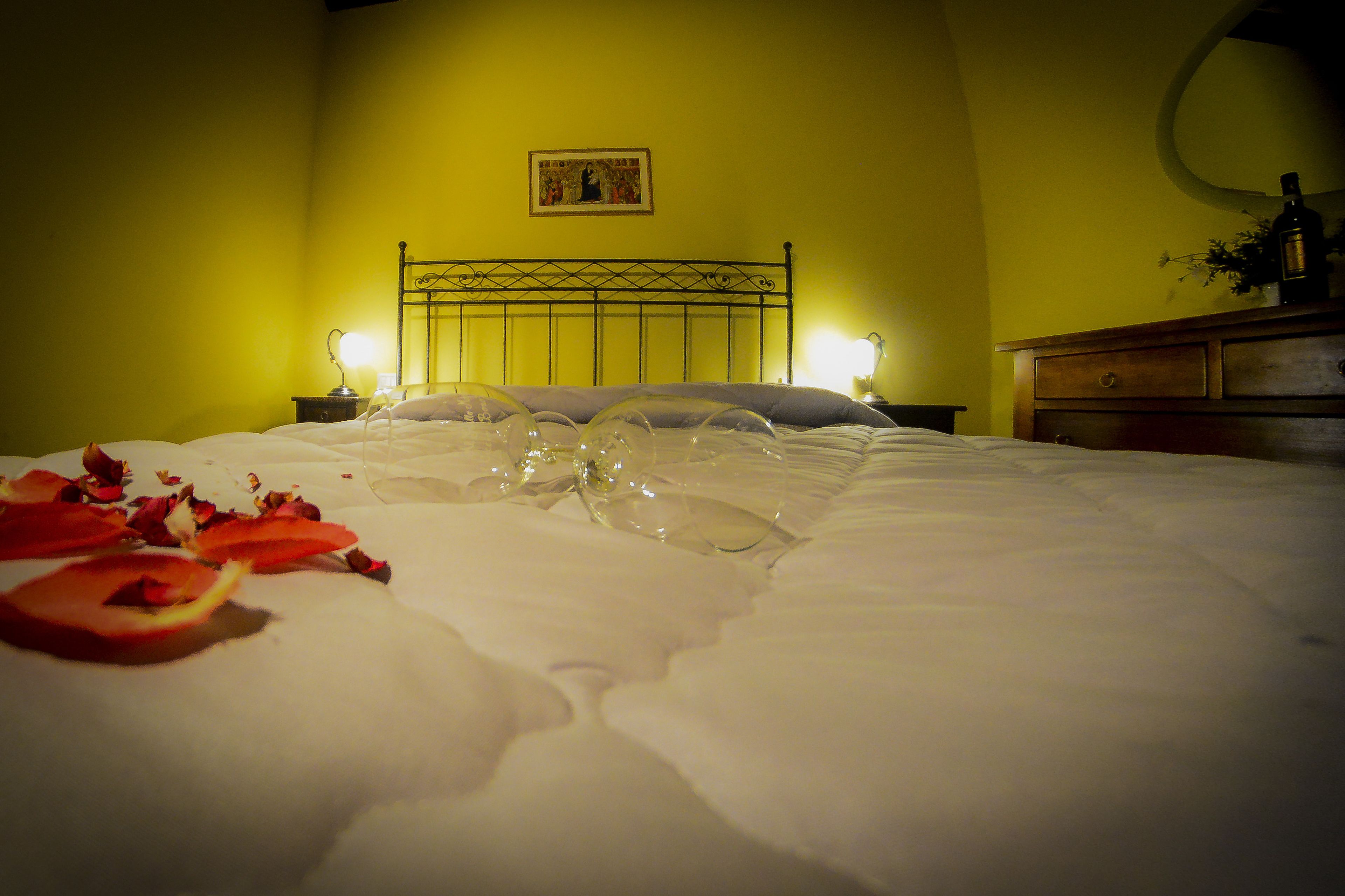 Una cama con pétalos de rosa y un cuenco de plástico transparente, una mesita de noche con una lámpara, y un aparador de madera con un espejo.