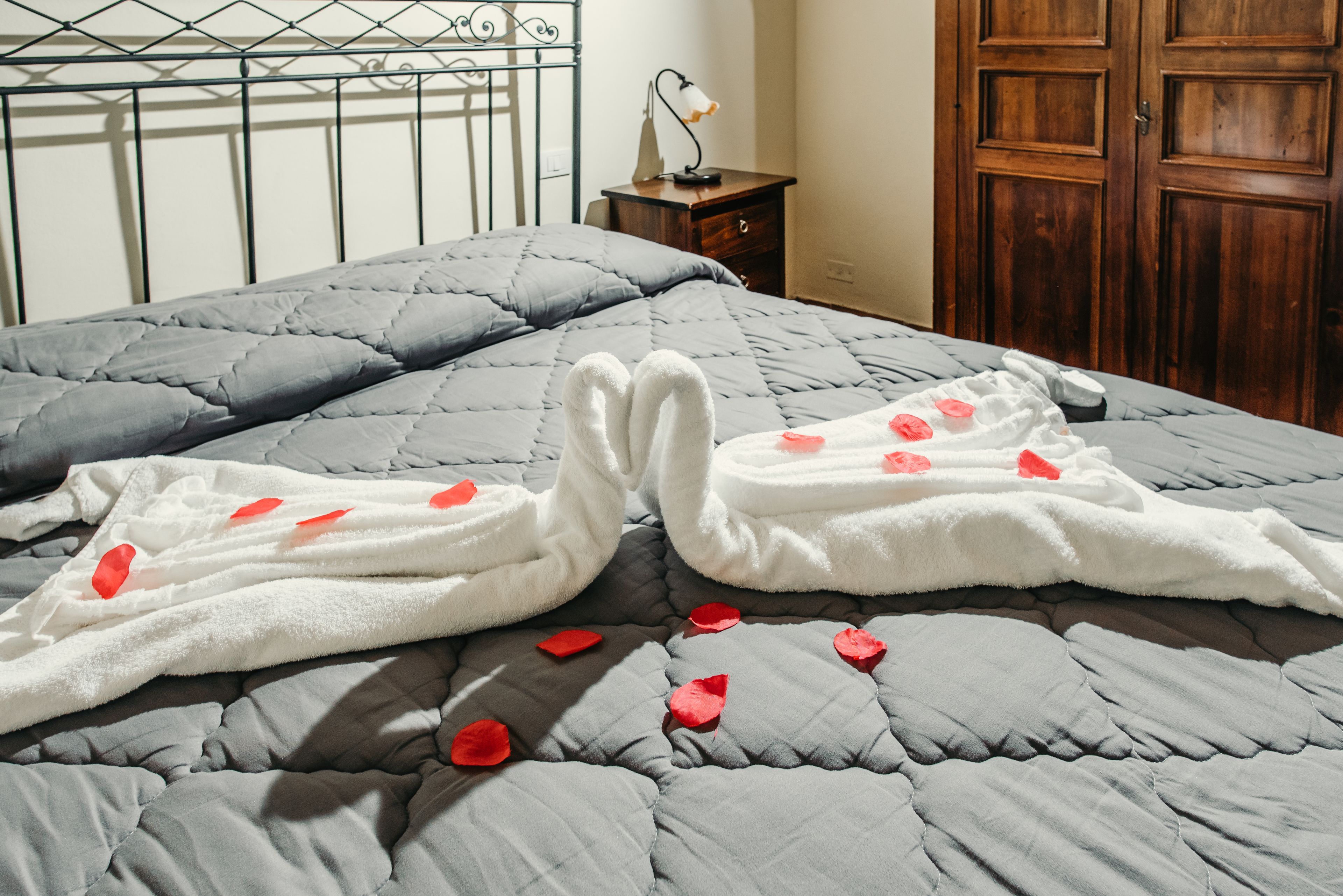 Een net opgemaakt bed met een grijze deken en witte handdoeken, met rozenblaadjes, een bedlampje en een houten garderobe in een hotelkamer.