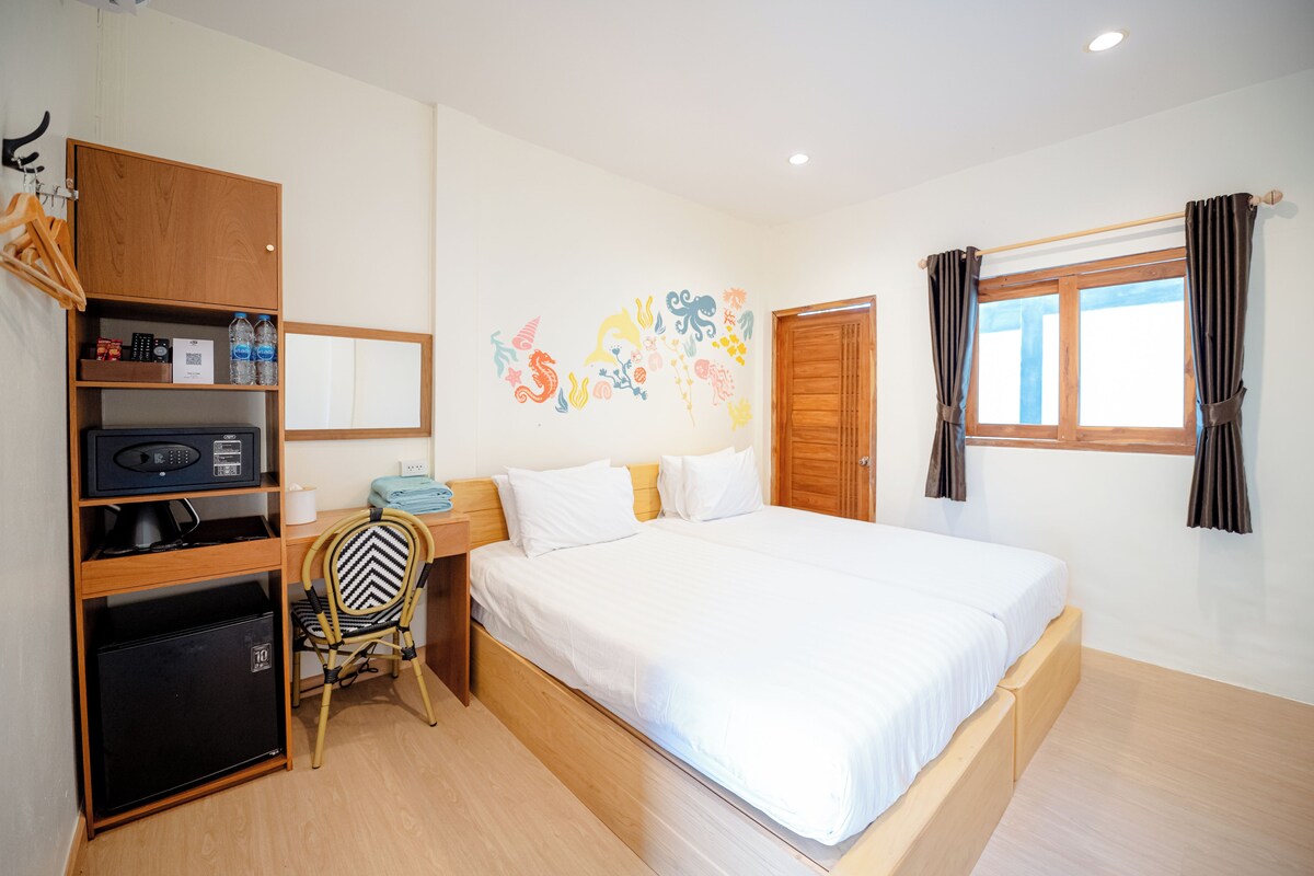 A hotel room with a bed, a desk with a chair, a wardrobe, a wall with a colorful mural, and a window with curtains.