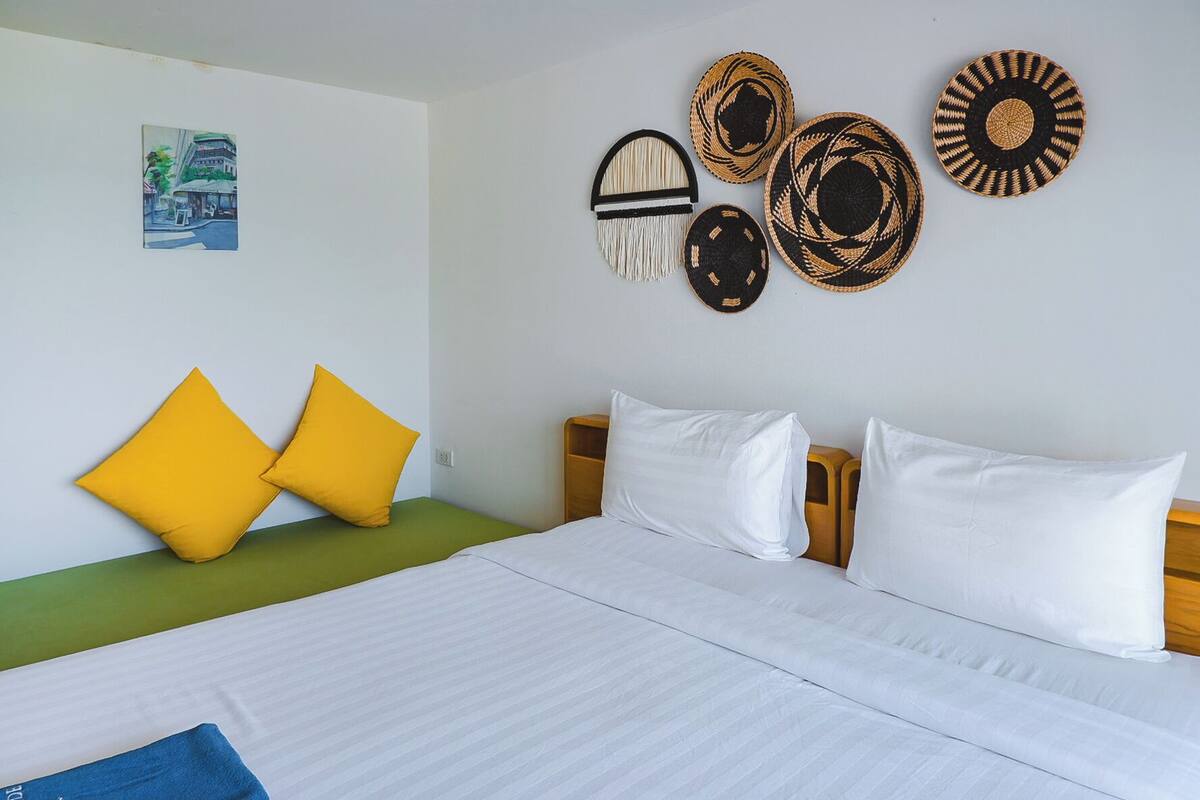 A hotel room with a bed, two yellow pillows, a green bench, and four woven wall hangings.