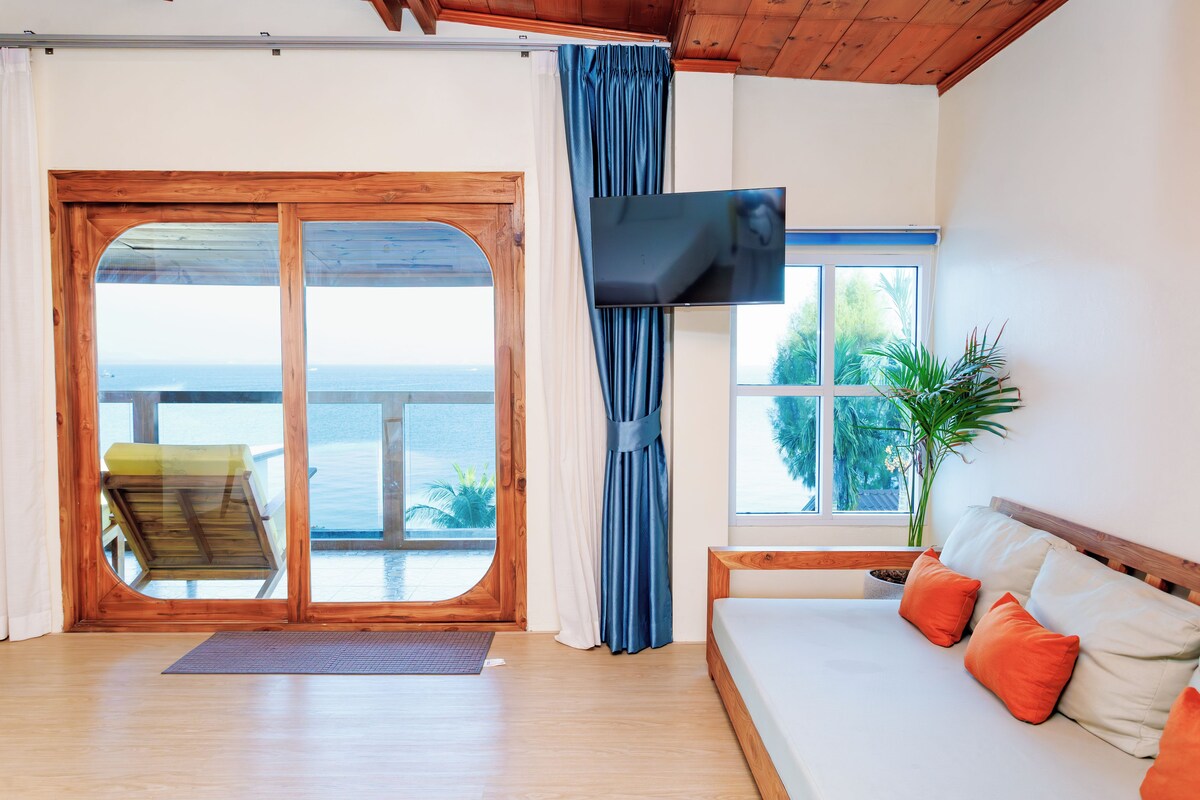 A room with a wooden floor, a sofa with orange cushions, a television, a large window with a view of the sea, and a sliding glass door leading outside.
