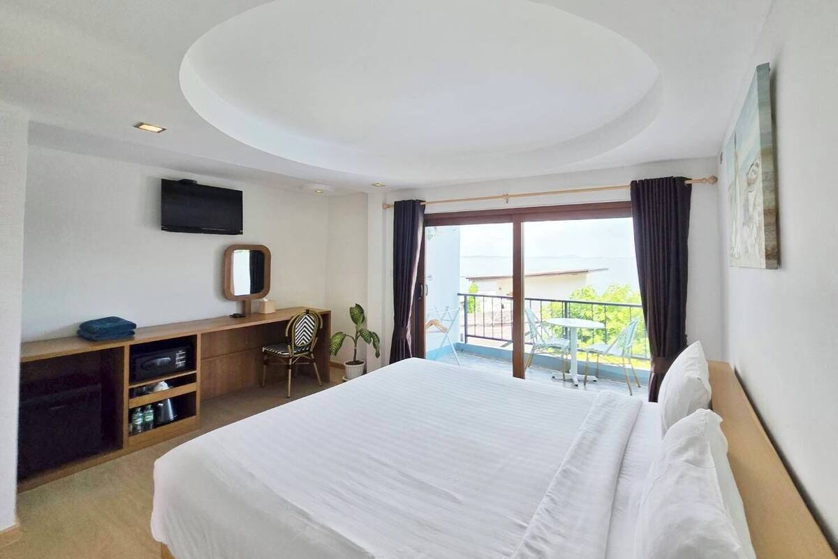 A hotel room with a bed, a desk, a chair, a TV, a balcony with a view, and a ceiling light.