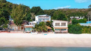 Una propiedad frente al mar con varias construcciones, incluyendo un edificio grande de color blanco con balcón y una estructura más pequeña con techo de paja.