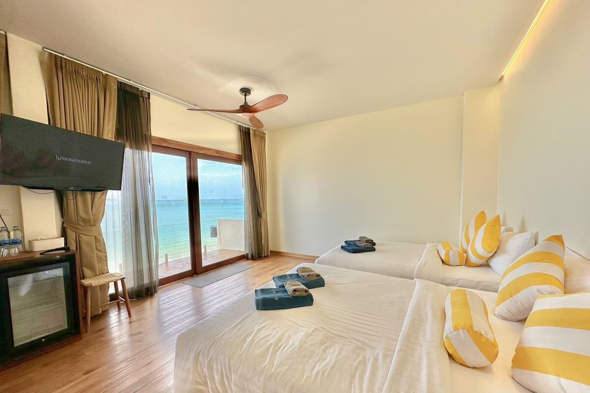 A hotel room with a TV, a bed with yellow and white striped pillows, a second bed, a ceiling fan, and a view of the sea through the windows.
