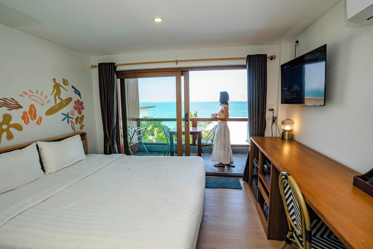 A hotel room with a bed, a desk, a TV, and a balcony with a view of the sea.