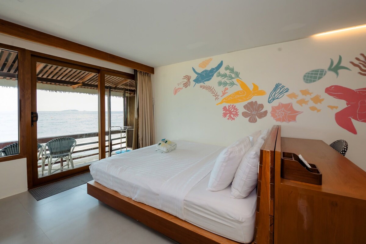 A hotel room with a bed, a wooden headboard, a balcony with chairs, and a wall with colorful marine life decals.