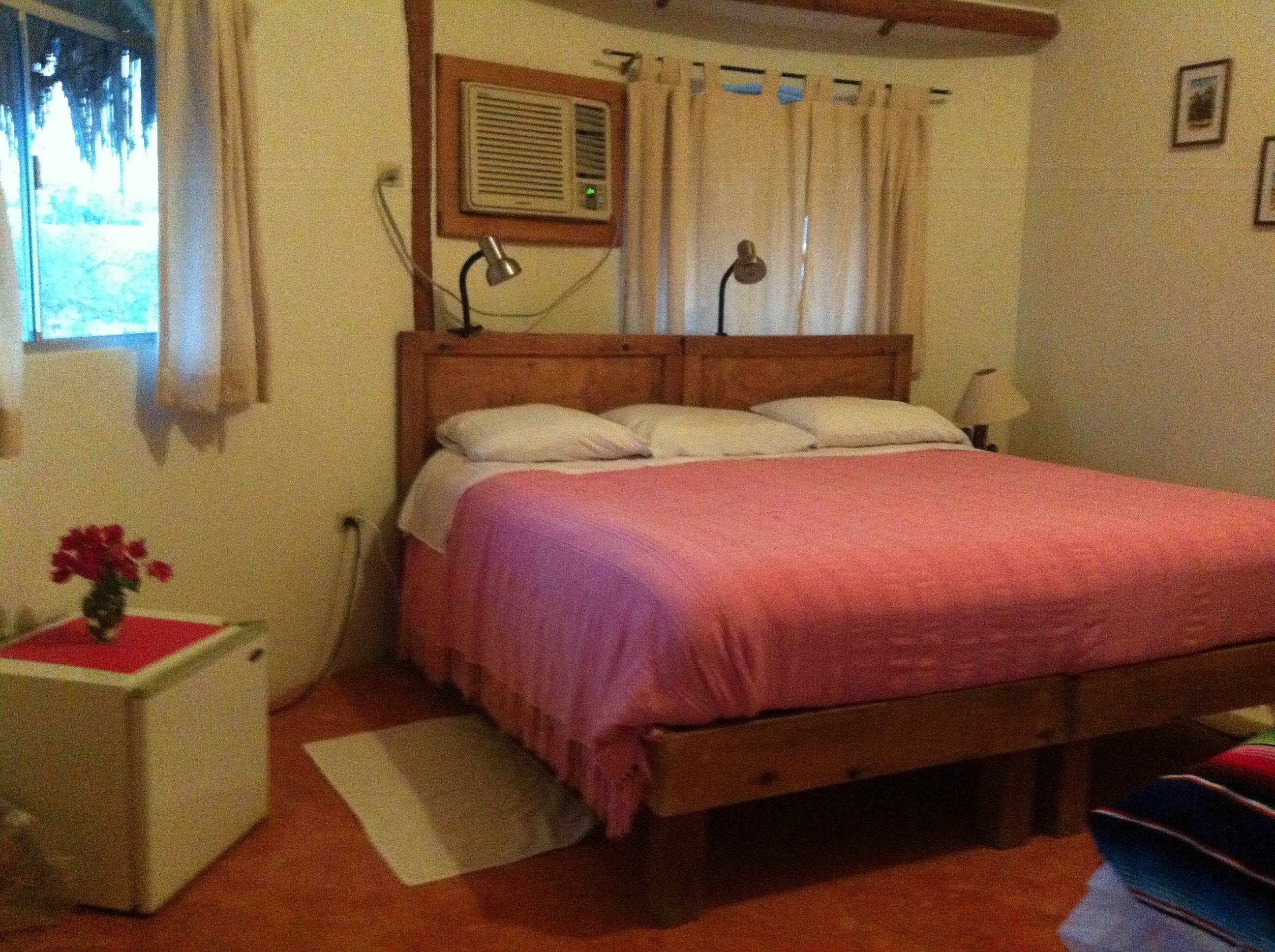 A bedroom with a wooden bed, white walls, a window with curtains, a bedside table with a lamp, and a small rug.