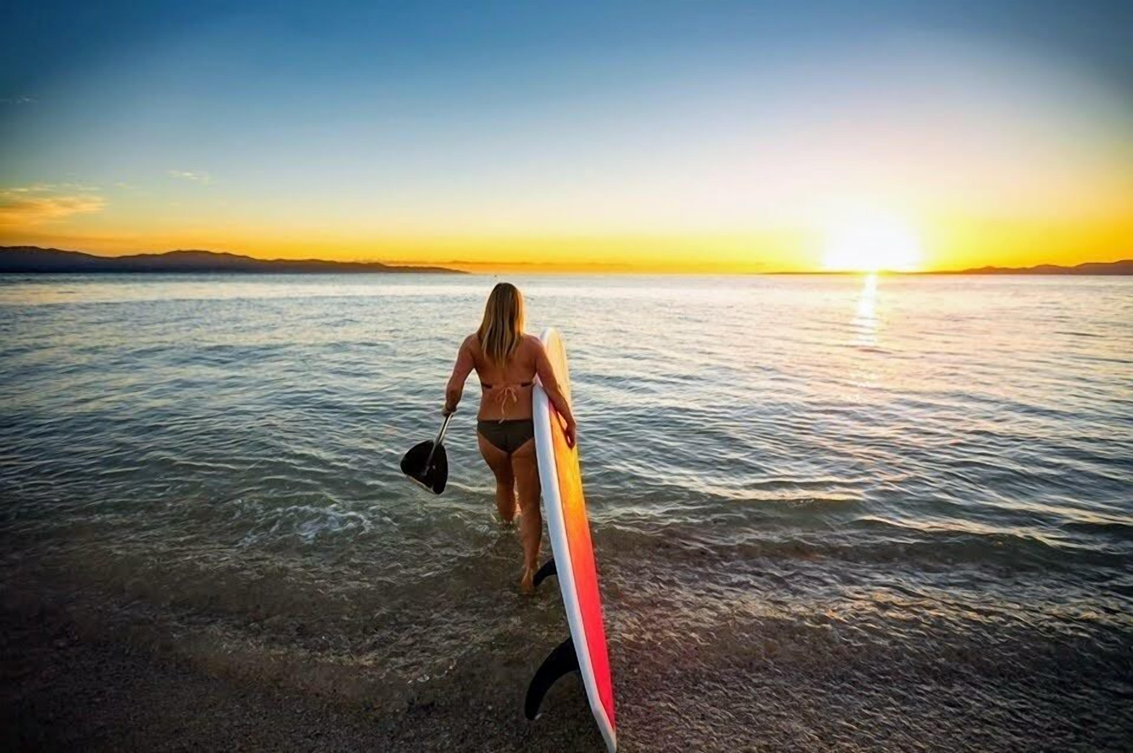 Una persona sosteniendo una tabla de surf en la playa al atardecer.