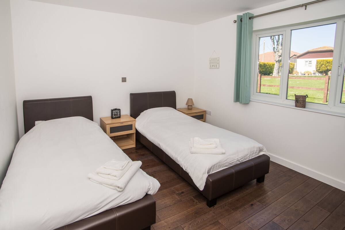 Quarto com duas camas, cabeceiras de madeira, mesa de cabeceira com abajur e janela com vista para uma casa e jardim.