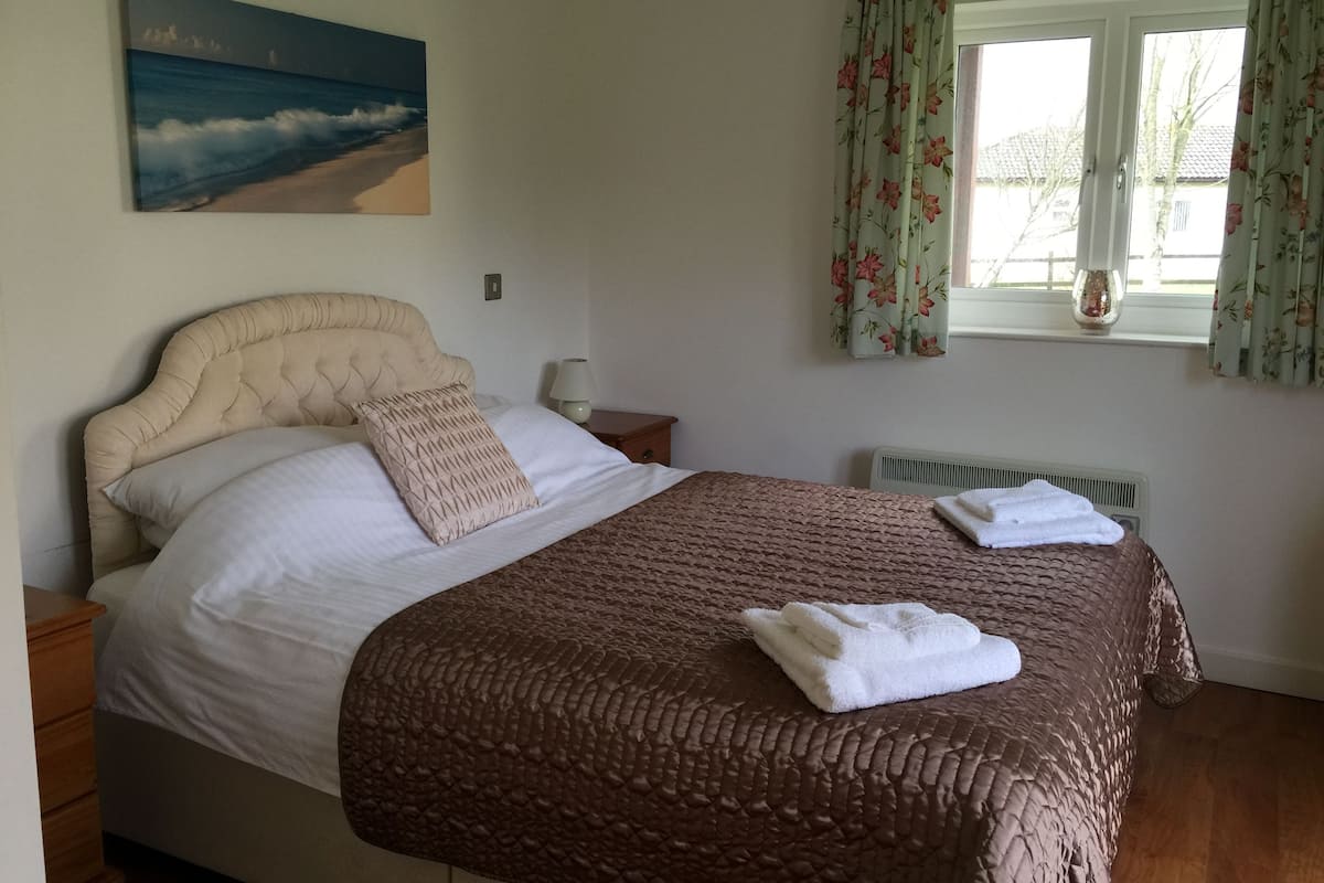 Cama bem arrumada com colcha marrom estampada e travesseiros, abajur, pintura de uma praia e janela com cortinas florais.