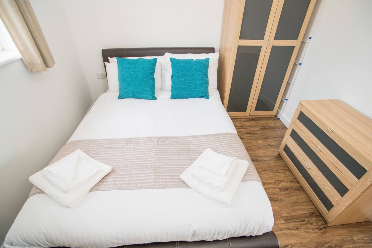 Uma cama bem arrumada com almofadas brancas e azul-turquesa, uma toalha branca dobrada e uma cômoda de madeira com gavetas.