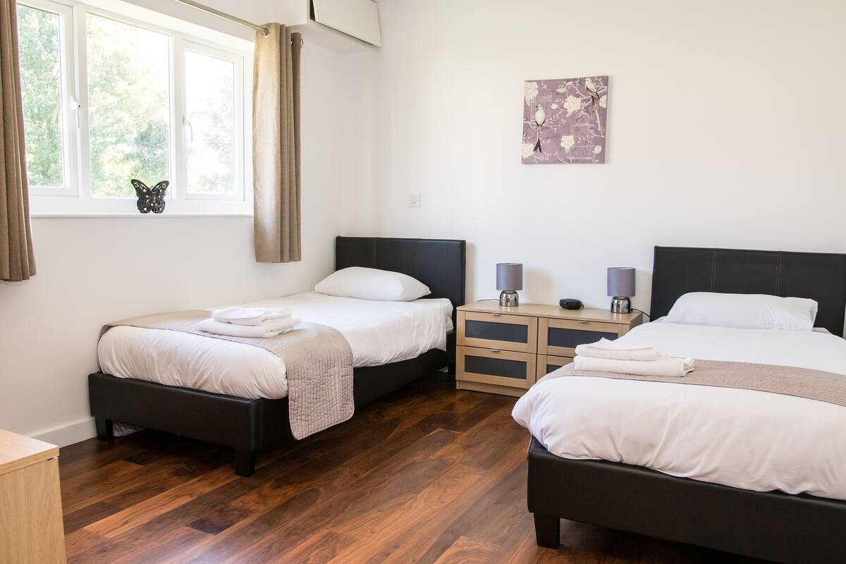 Quarto de hotel com duas camas, piso de madeira, mesa de cabeceira com luminárias, janela com cortinas e uma obra de arte na parede.