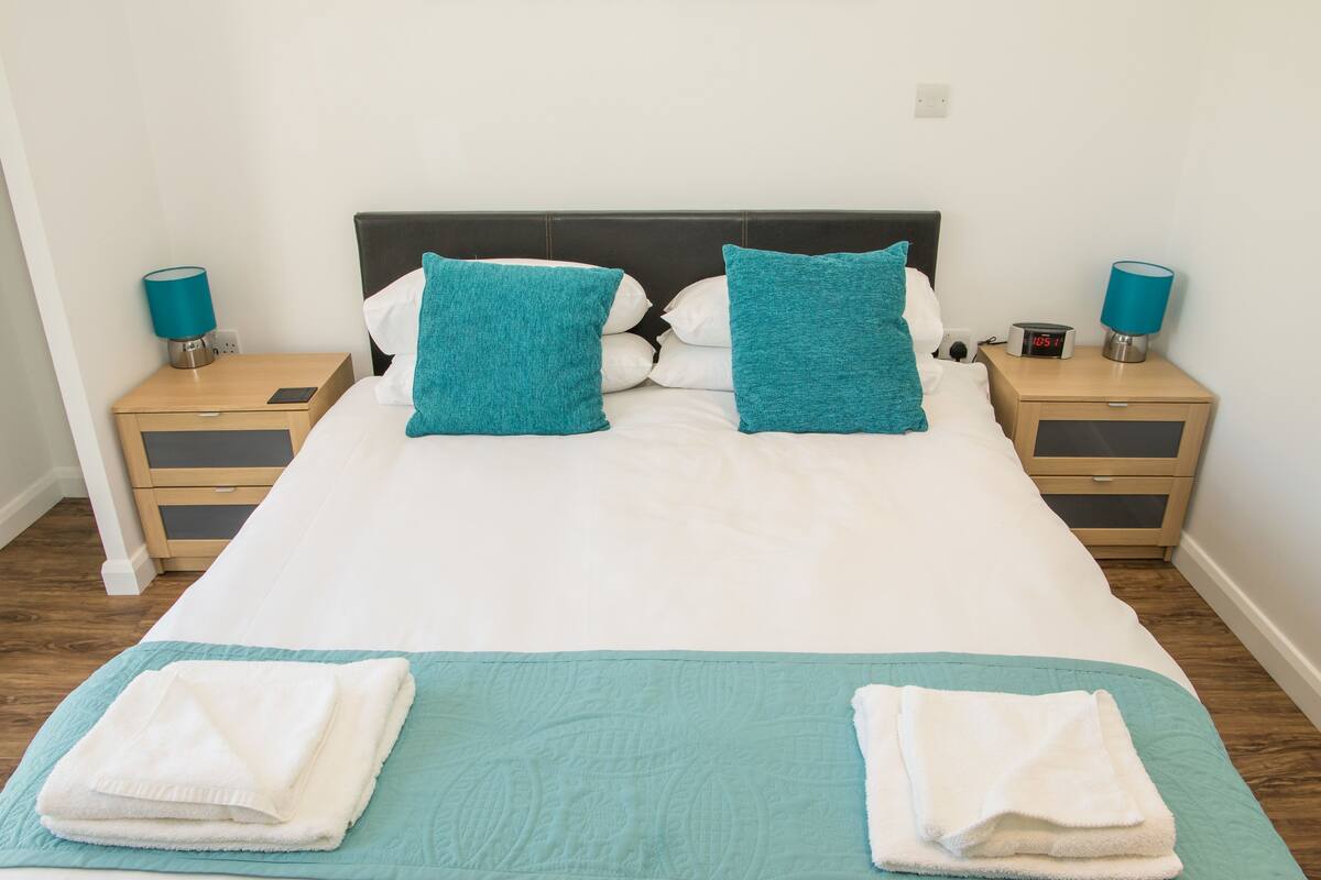 Cama bem arrumada com roupa de cama branca e azul-turquesa, dois travesseiros azul-turquesa e um cobertor azul-turquesa. Há duas mesas de cabeceira com abajures e um pequeno relógio.