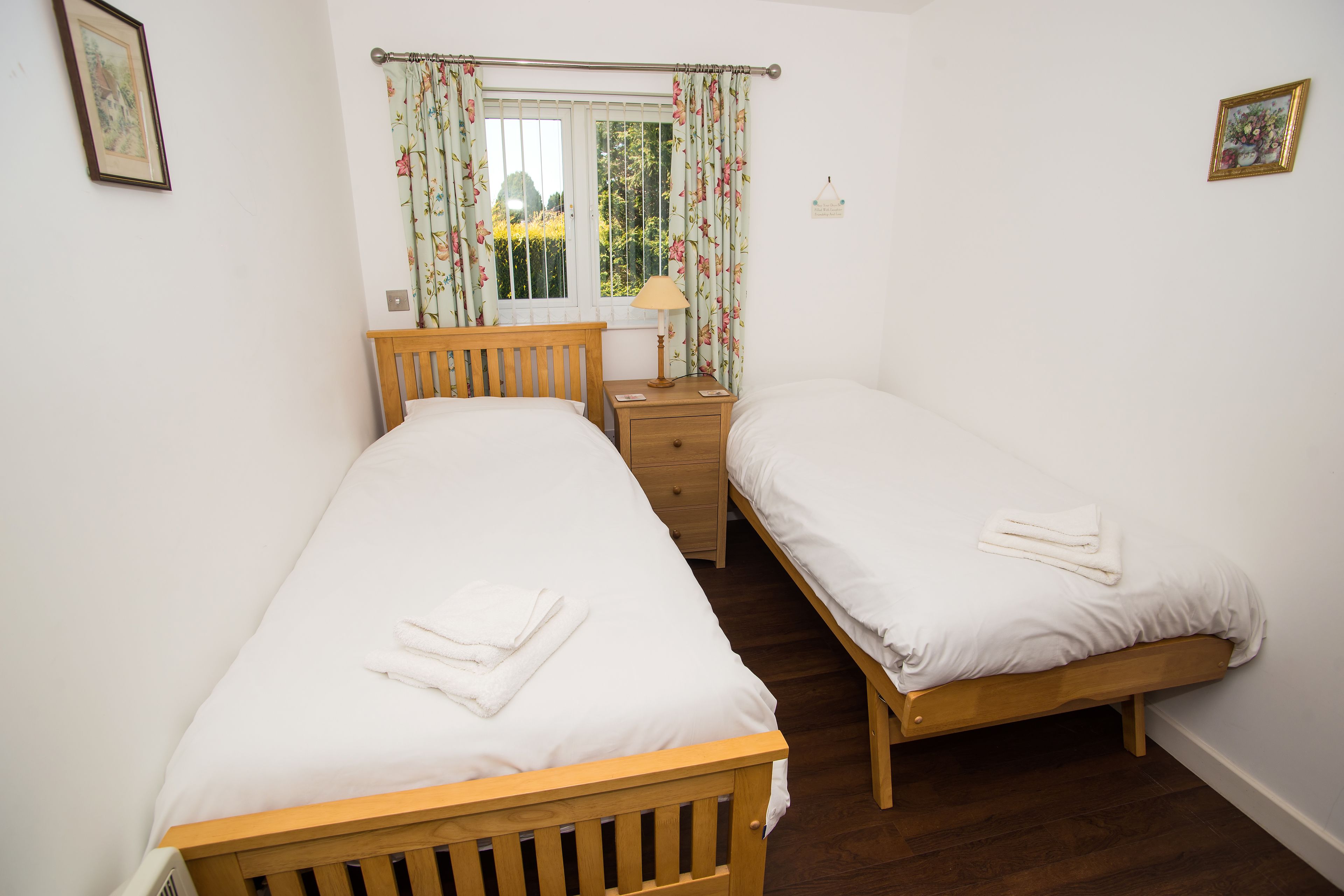 Quarto com duas camas de solteiro, uma mesinha de cabeceira, uma janela com cortinas florais e uma pequena mesa ao lado da cama.
