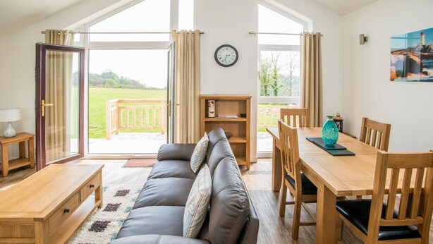 A cozy living room with a sofa, wooden dining table, and chairs, a bookshelf, and a large window with curtains. - Stoneleigh Village (Sidmouth)