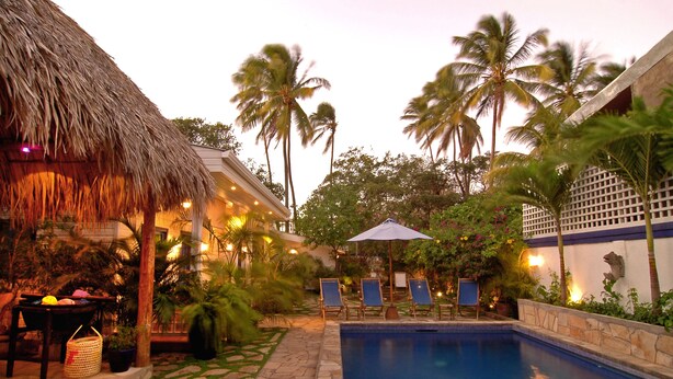 A pool area with lounge chairs, a thatched-roof structure, and palm trees. - La Posada Azul (San Juan del Sur)