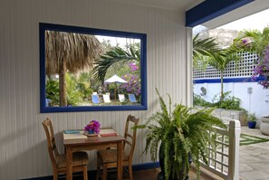 Uma sala de jantar com mesa, cadeiras e janela com vista para um jardim com plantas e uma estrutura com telhado de palha.