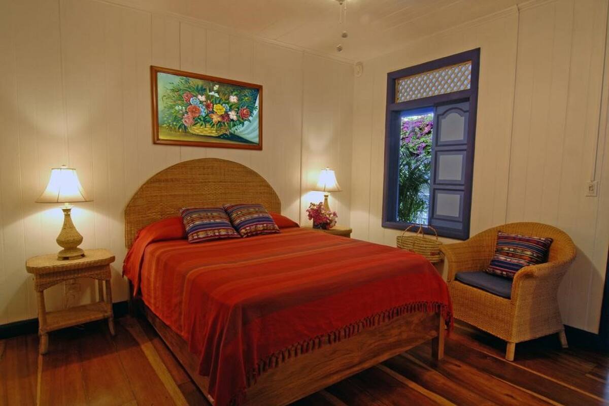 A bedroom with a wooden bed, two bedside tables with lamps, a wicker chair, and a window with a view of greenery.