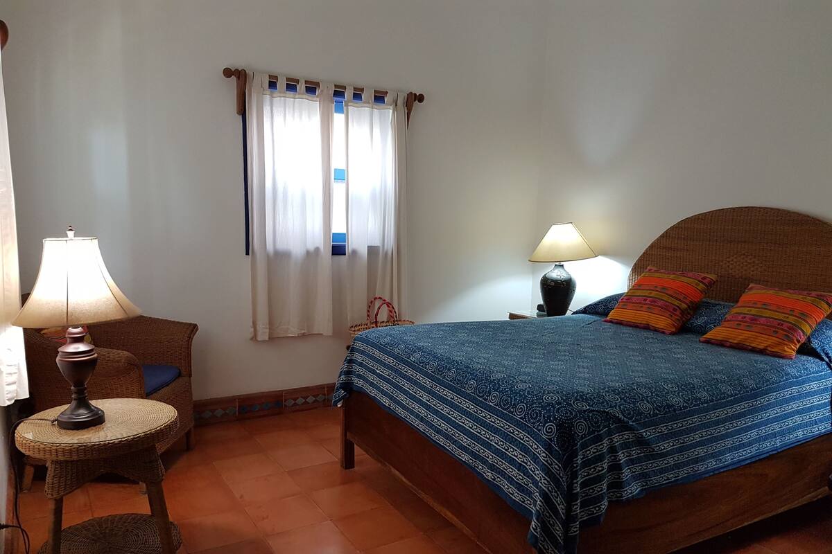 A bedroom with a bed, two bedside lamps, a wicker chair, a small table, and a window with curtains.