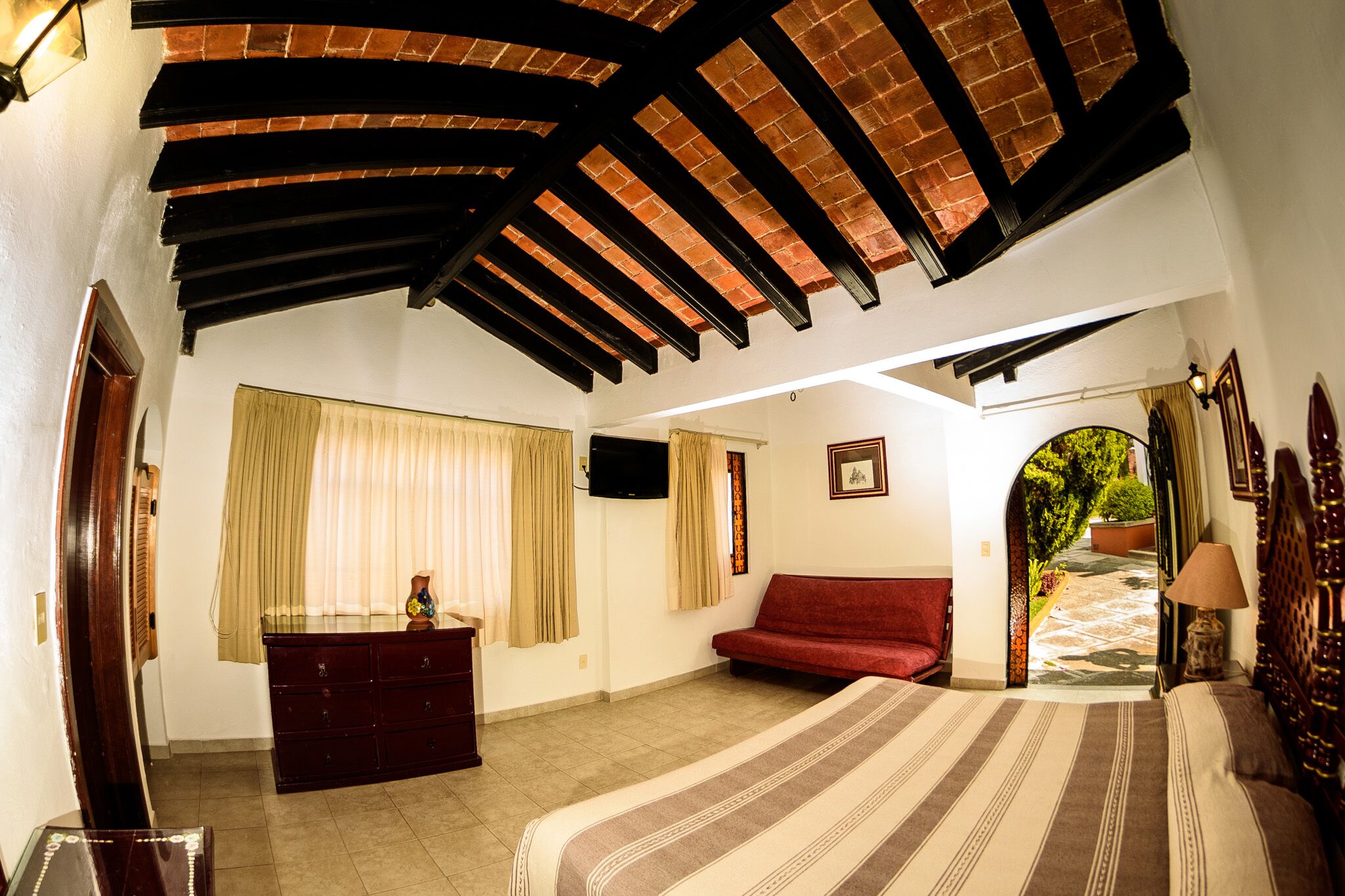 Un dormitorio con cama, sofá rojo, cómoda y televisor. Hay una ventana con cortinas y una puerta que da a un balcón.