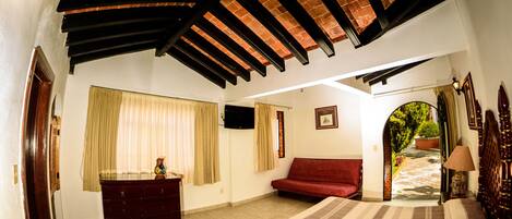 Un dormitorio con cama, sofá rojo, cómoda y televisor. Hay una ventana con cortinas y una puerta que da a un balcón.