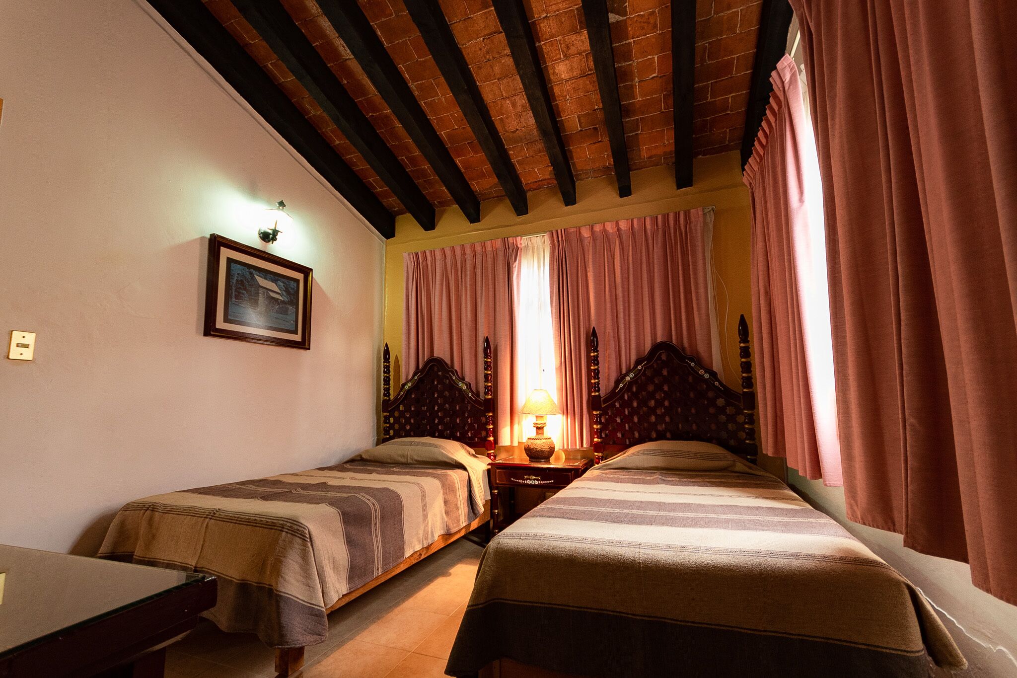 Una habitación con dos camas individuales, una mesa de madera, un cuadro en la pared y una ventana con cortinas rosadas.