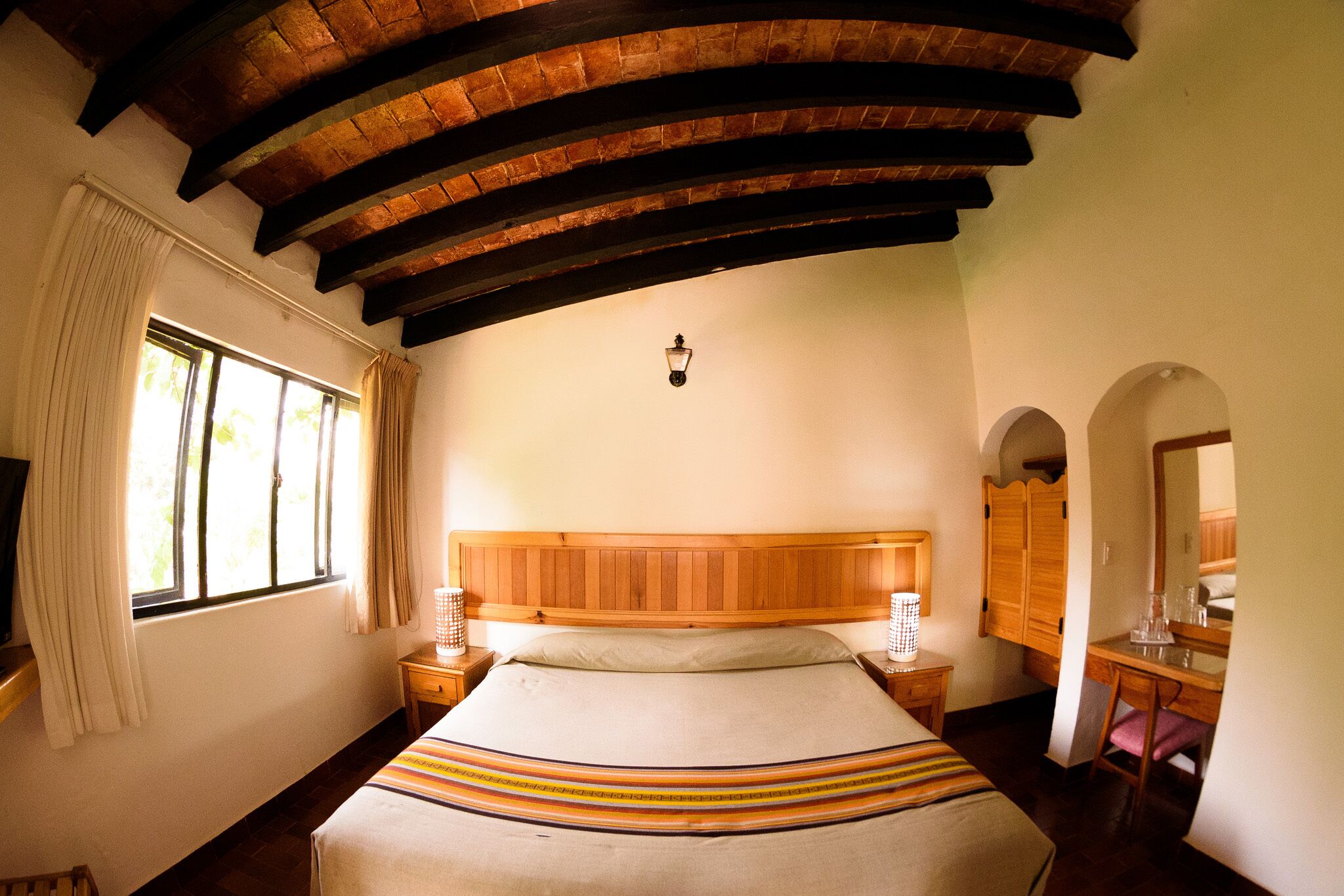 Una camera da letto con testiera in legno, un letto con copripiumino a righe, un comodino con lampada, una finestra con tende e un'armadio in legno.